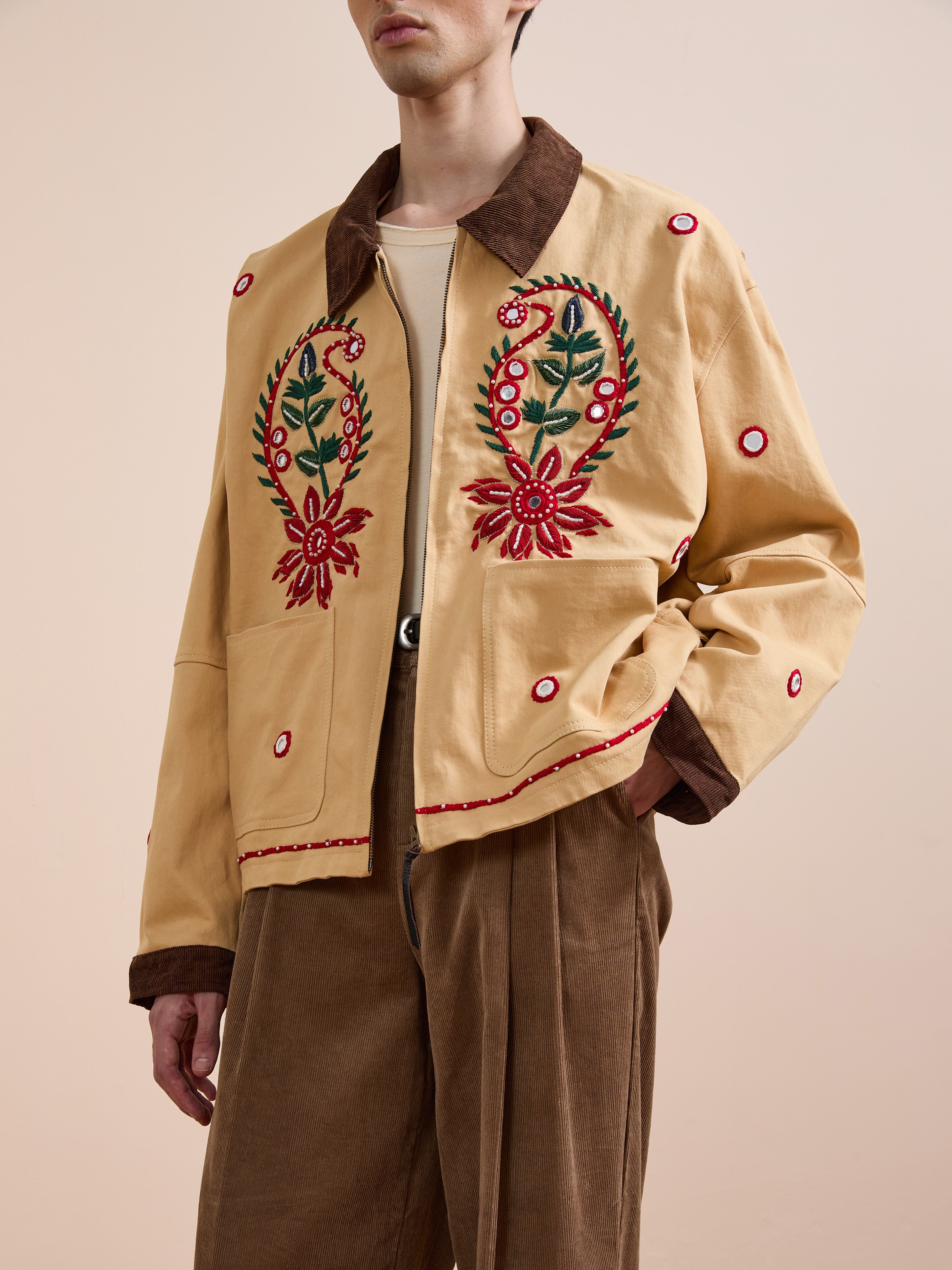 A person models the Found Paisley Motif Hand Embroidered Work Jacket, featuring red and green accents, a brown collar, and artisan craftsmanship, styled with brown trousers against a plain background.