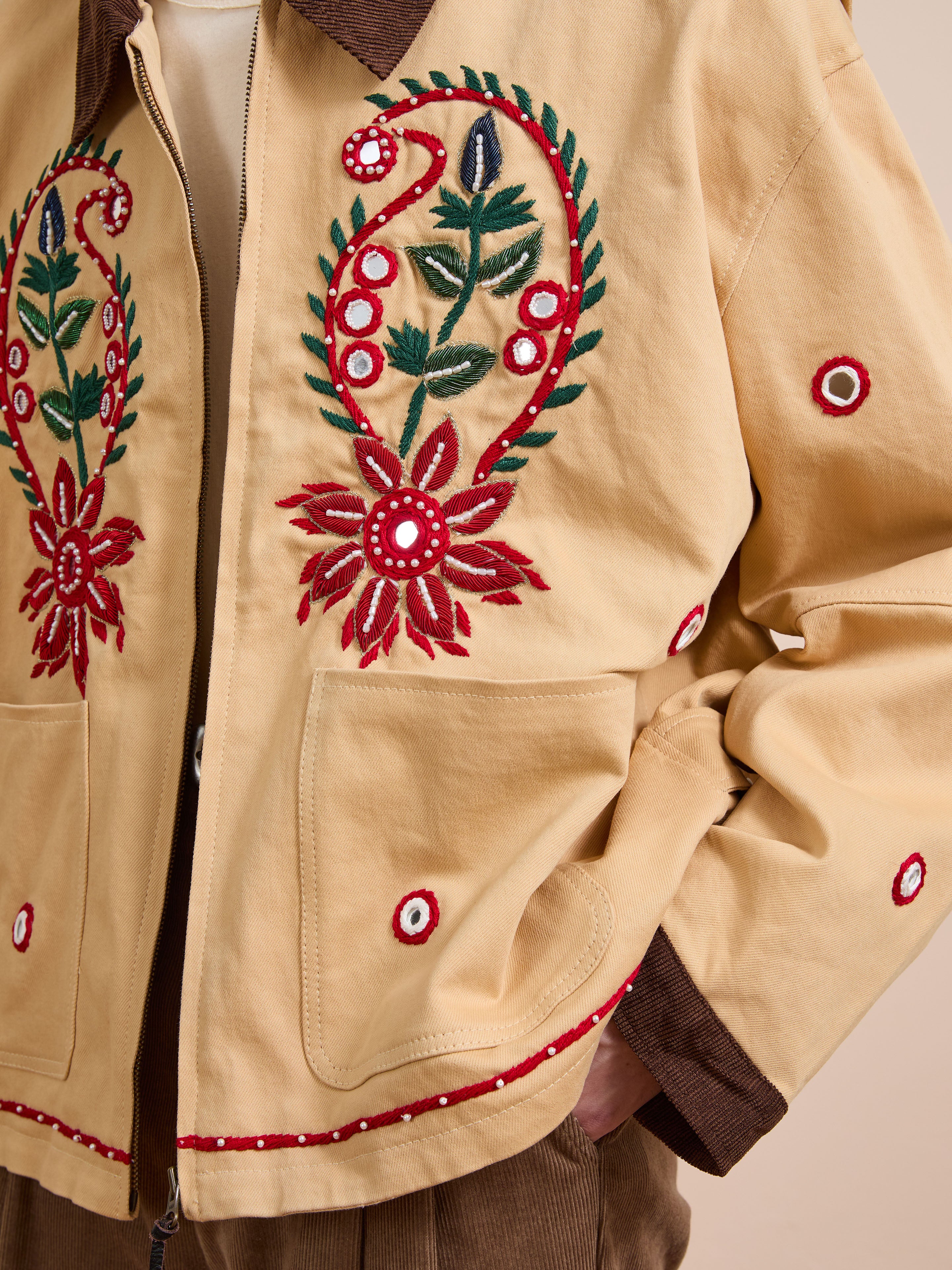 A person wearing the Found Paisley Motif Hand Embroidered Work Jacket in tan, featuring red and green paisley floral embroidery with circular patterns, shown from shoulders to waist.