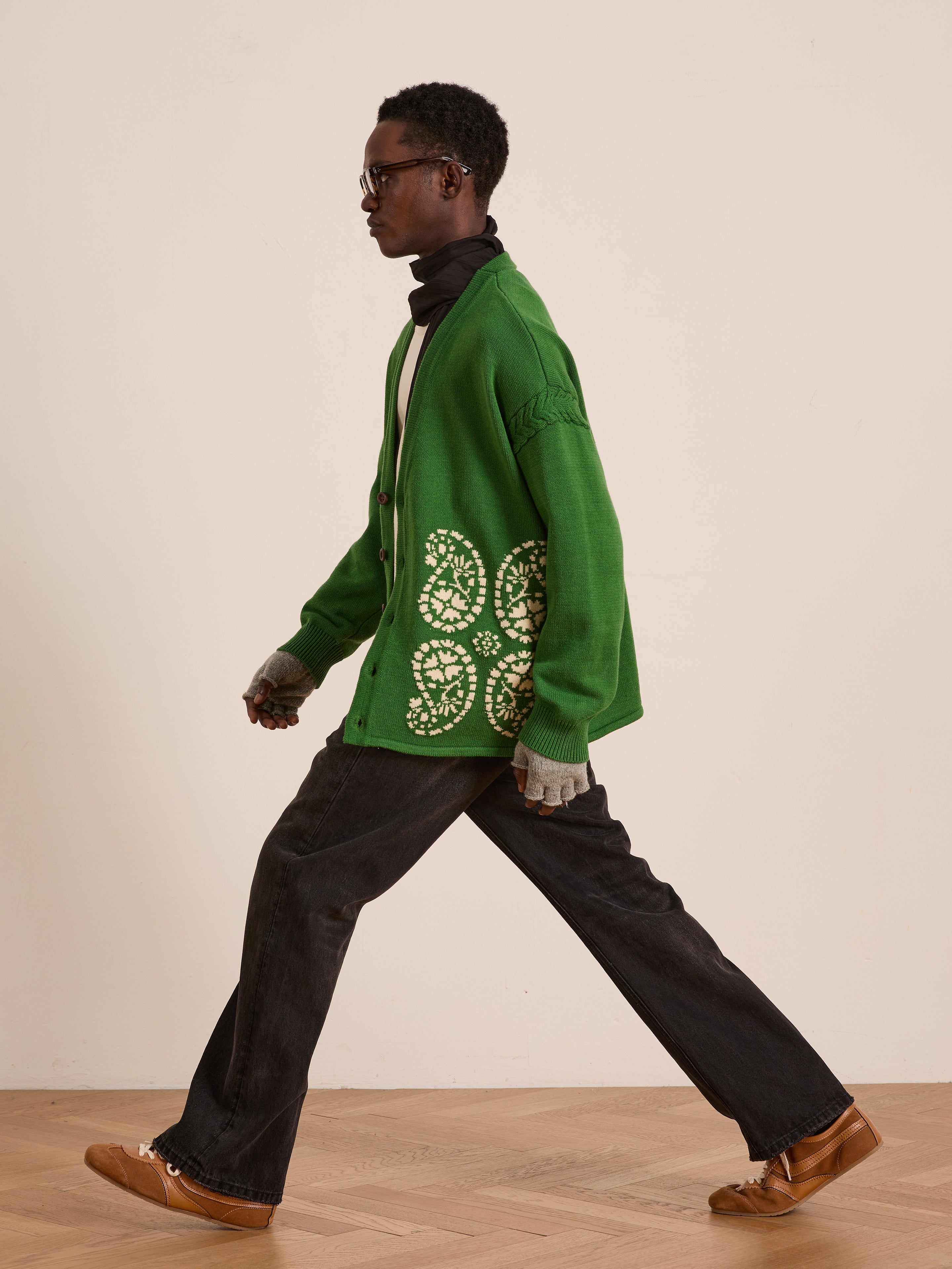 Wearing the FOUND Paisley Knit Cardigan, a person in glasses walks indoors on a wooden floor against a plain wall, paired with black pants and brown shoes.