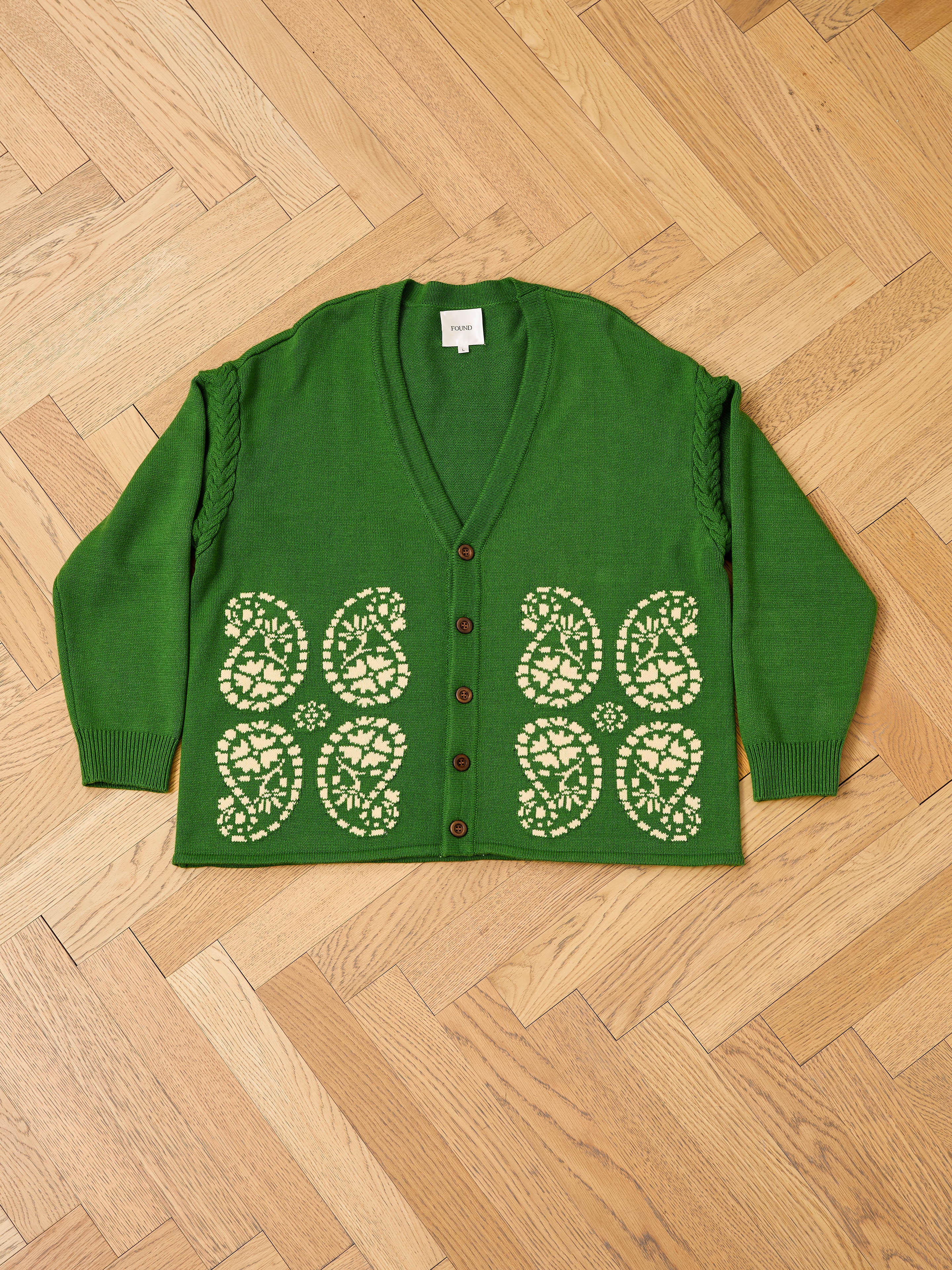 The FOUND Paisley Knit Cardigan is a green unisex V-neck button-up, featuring white floral and South Asian-inspired patterns on the lower front, shown laid flat on a wooden parquet floor.