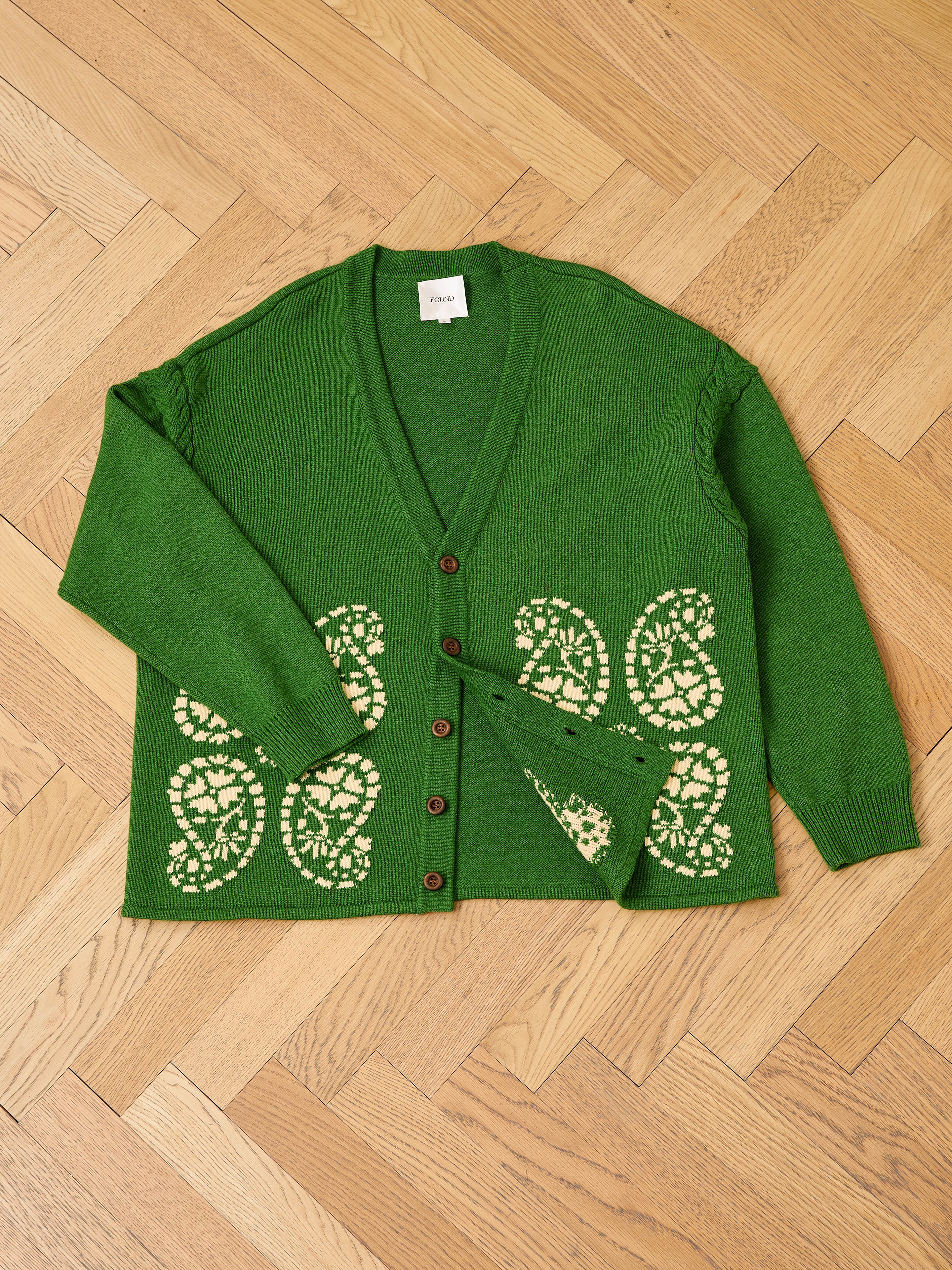 The FOUND Paisley Knit Cardigan, a unisex green cotton knit with brown buttons and cream patterns, is displayed flat on a wooden herringbone floor.