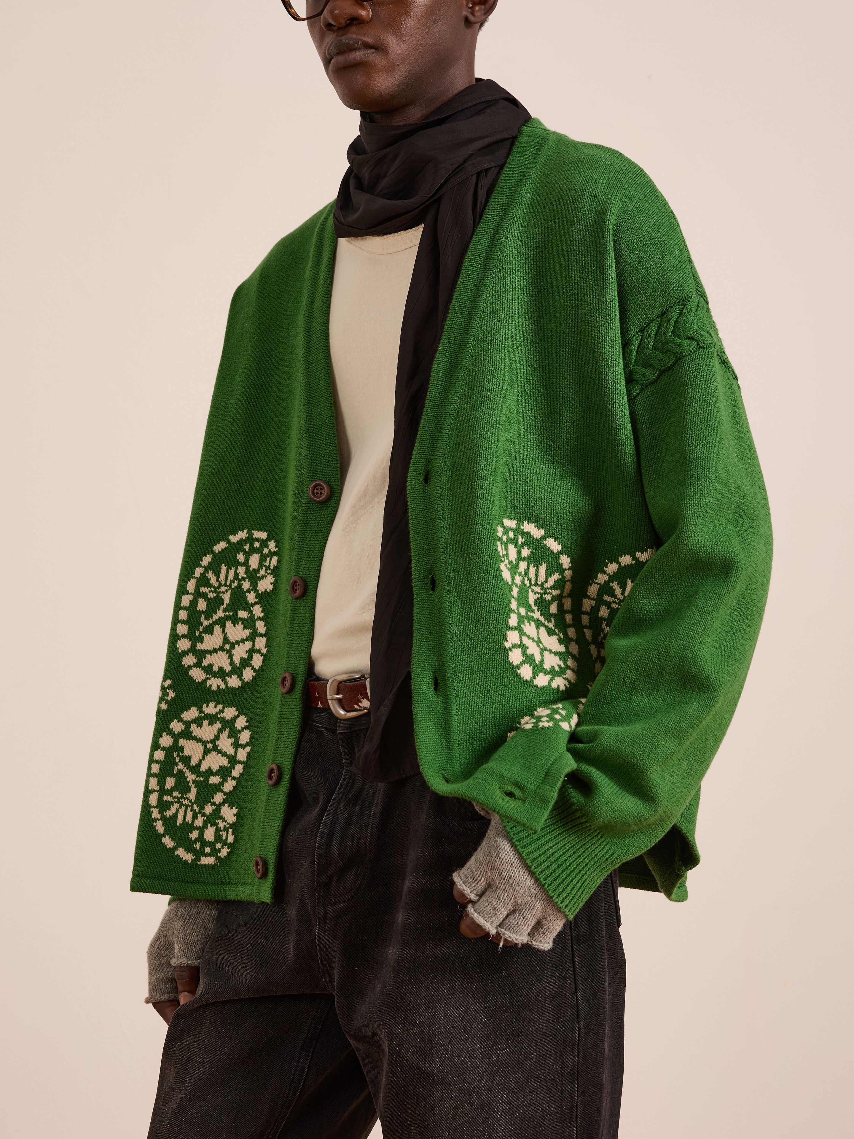 A person wearing the FOUND Paisley Knit Cardigan in green with white patterns, paired with a cream shirt, black scarf, gray fingerless gloves, dark jeans, and glasses stands against a plain background.