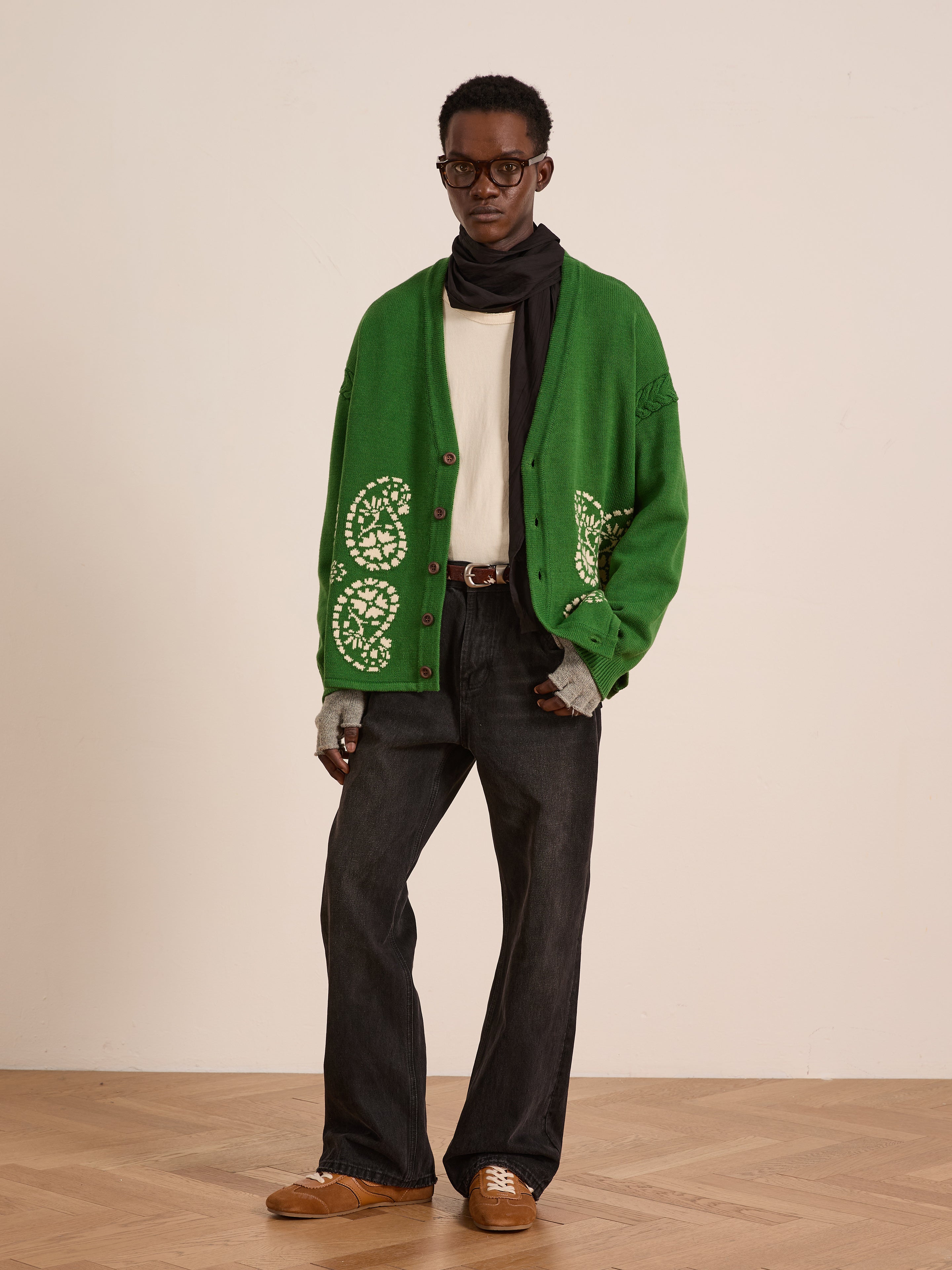 A man stands indoors wearing the FOUND Paisley Knit Cardigan in green with white patterns, paired with a white shirt, dark jeans, brown shoes, a black scarf, glasses, and gray gloves.