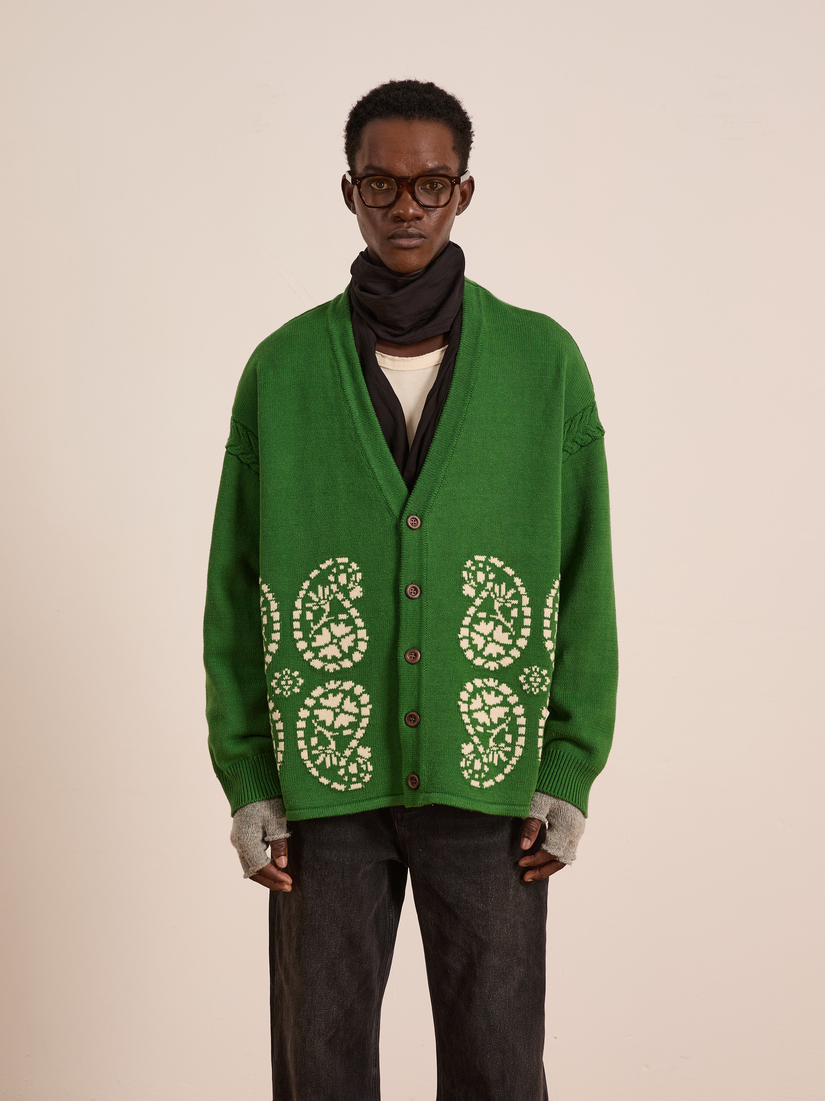 A person wearing glasses, a black scarf, gray fingerless gloves, a white shirt, dark jeans, and the FOUND Paisley Knit Cardigan with green and white South Asian patterns stands against a plain background.