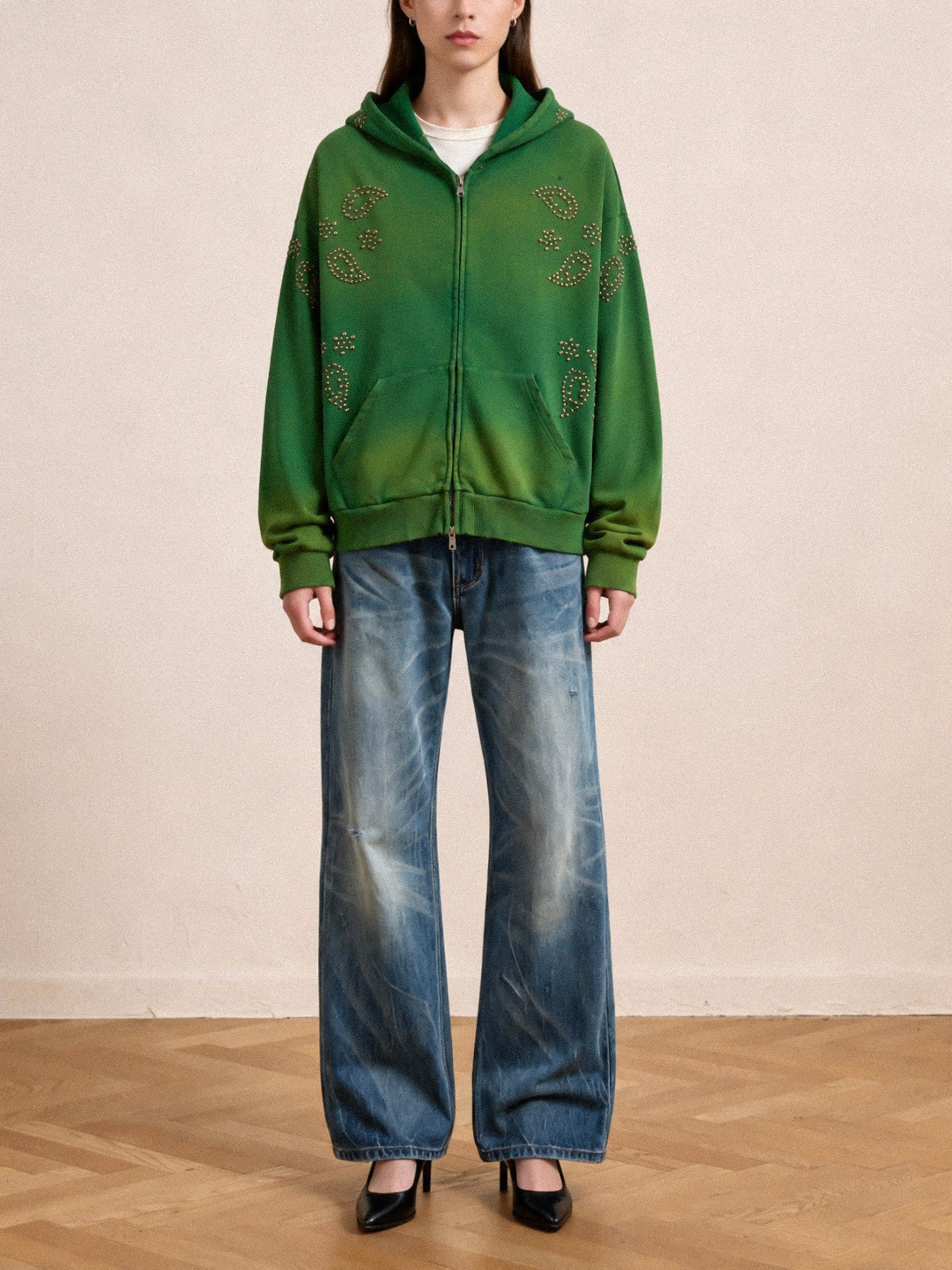A person models the FOUND Paisley Studded Zip Up Hoodie featuring star patterns, styled with blue wide-leg jeans and black heels, standing on a wooden floor against a plain background.