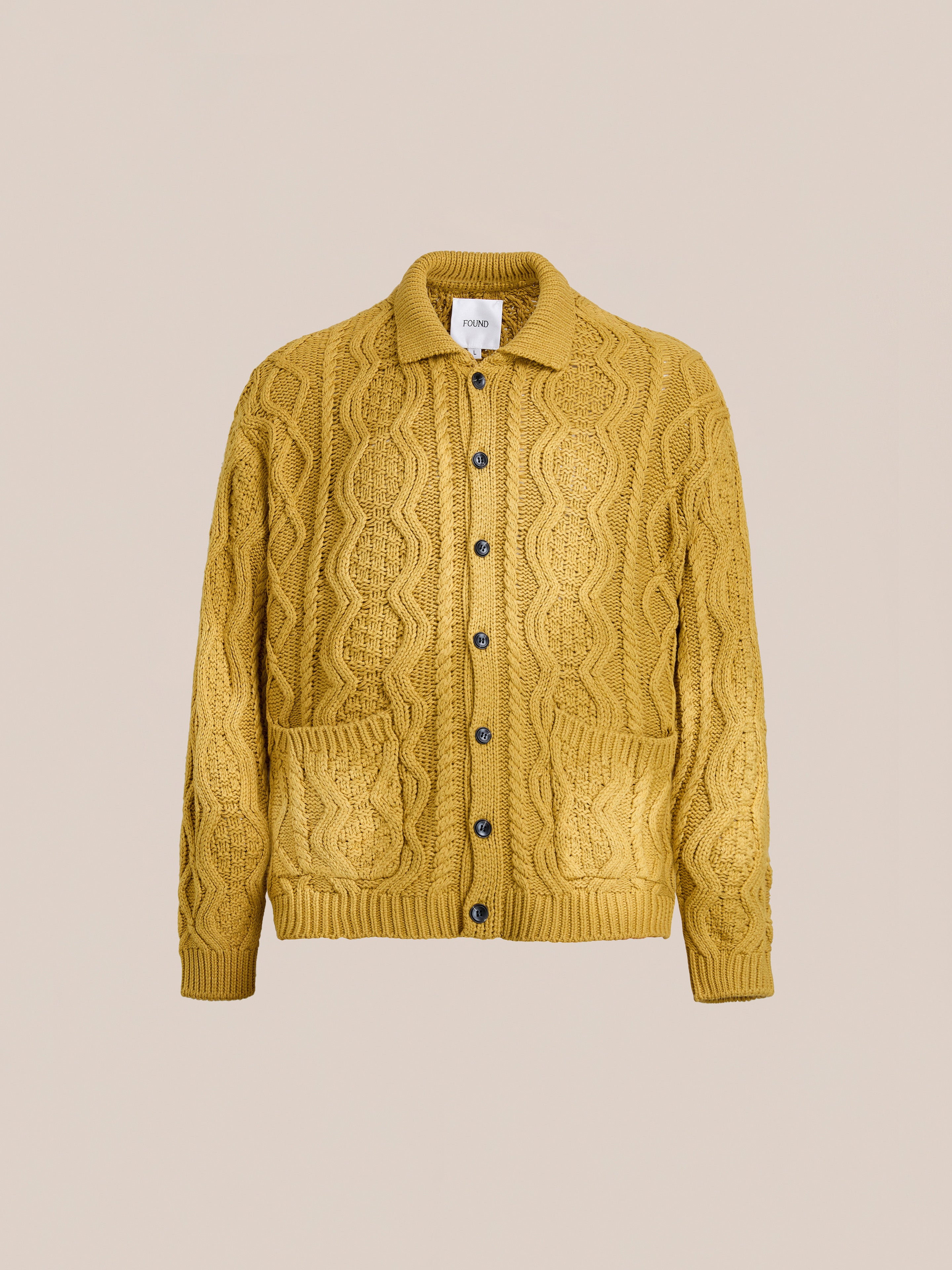 The FOUND Cable Knit Buttoned Cardigan is a unisex mustard yellow sweater featuring chunky cables, front buttons, two pockets, and a collar, shown on a plain beige background.