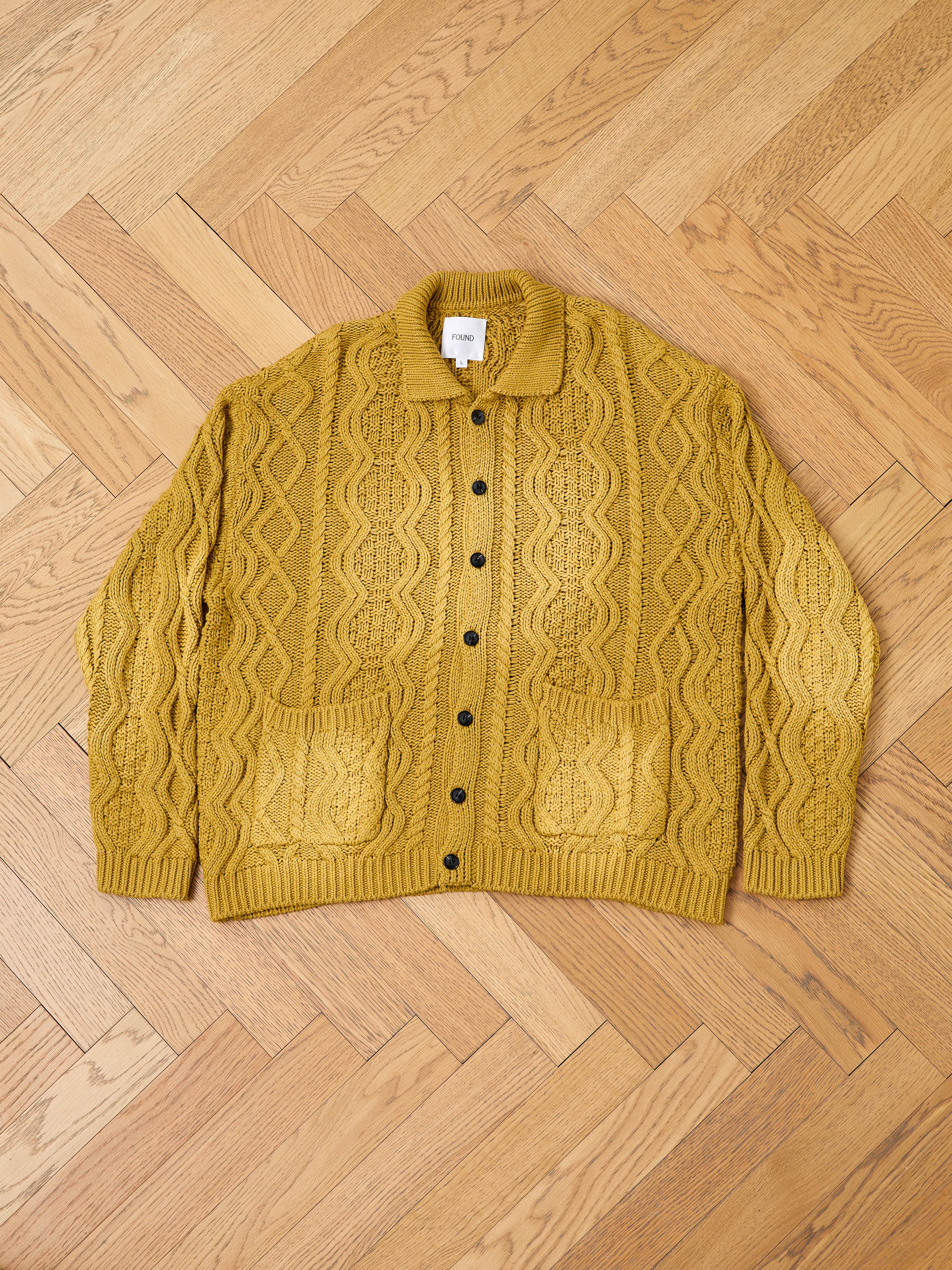 The FOUND Cable Knit Buttoned Cardigan in mustard yellow features chunky cables, black buttons, and two front pockets, shown laid flat on a light wood herringbone floor.