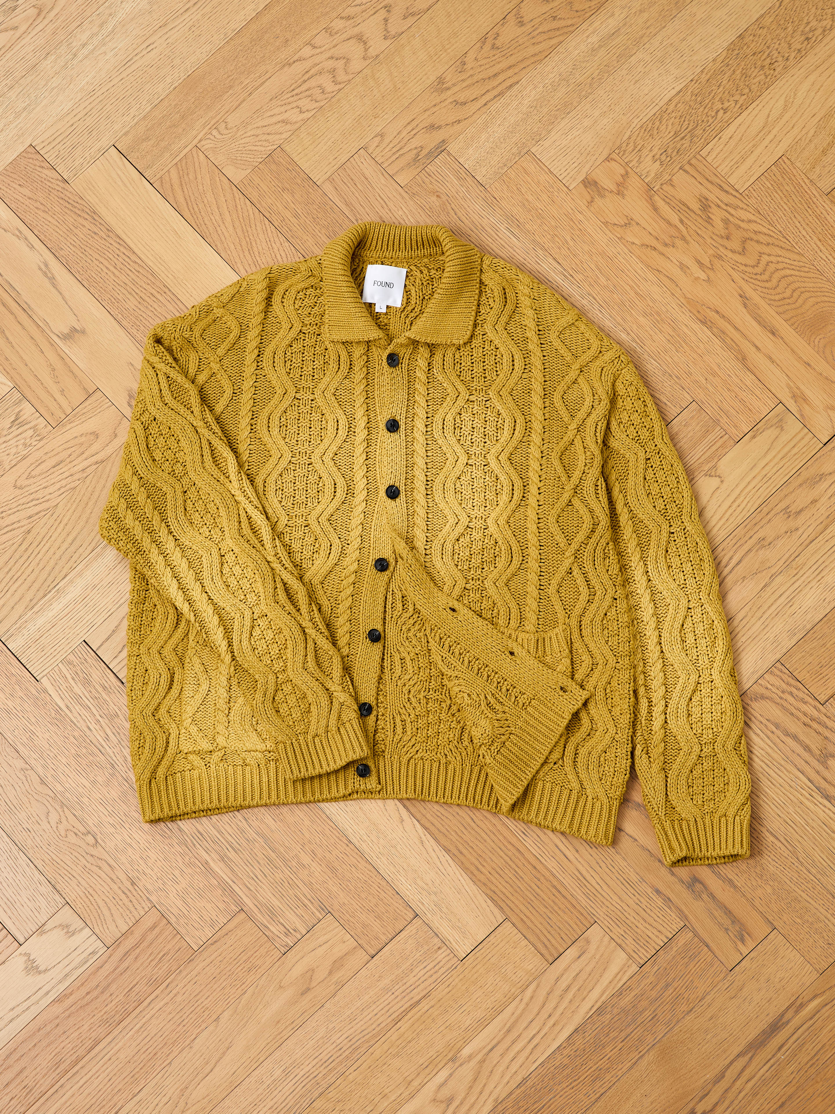 The FOUND Cable Knit Buttoned Cardigan in mustard yellow, featuring chunky cable knitting and black buttons, is displayed flat on a light wooden floor.