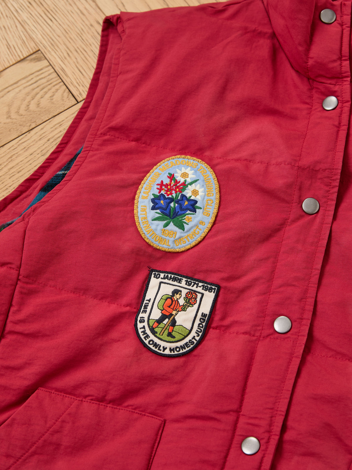 FOUND Patch Down Vest in red features two embroidered chest patches: one round with text and florals, the other rectangular with a person. Shown on a wooden floor.