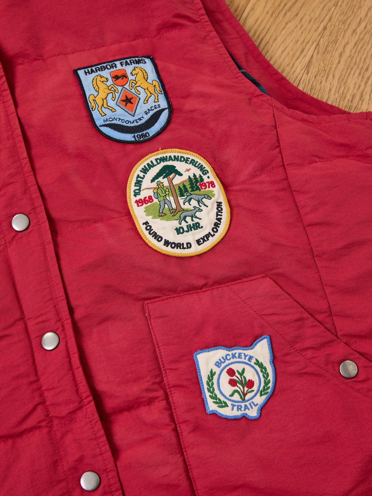 FOUND Patch Down Vest: Red outdoor vest with silver snap buttons and three colorful patches—Harbor Farms, Buckeye Trail, and round world event badges.