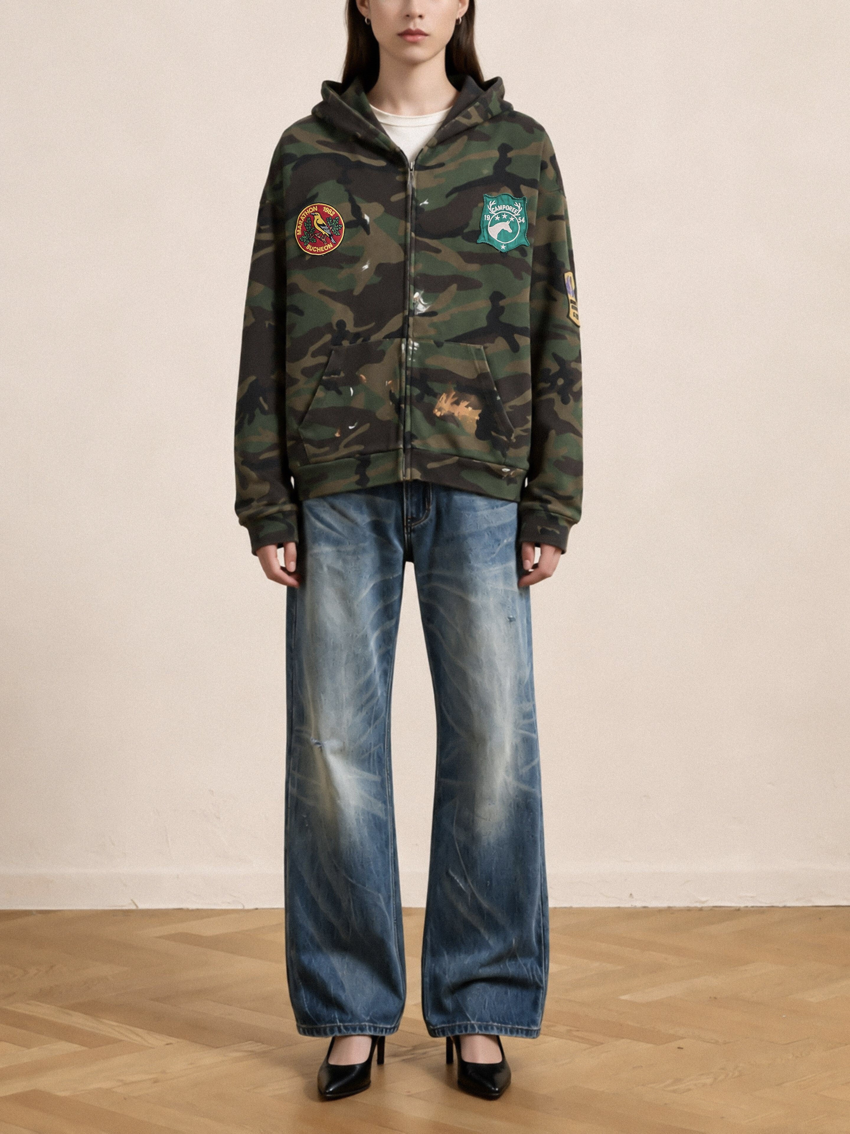 A person stands against a plain wall, facing forward with hands by their sides, wearing the FOUND Patch Camo Zip Up Hoodie styled with unisex relaxed fit, wide-leg faded jeans and black pointed heels.