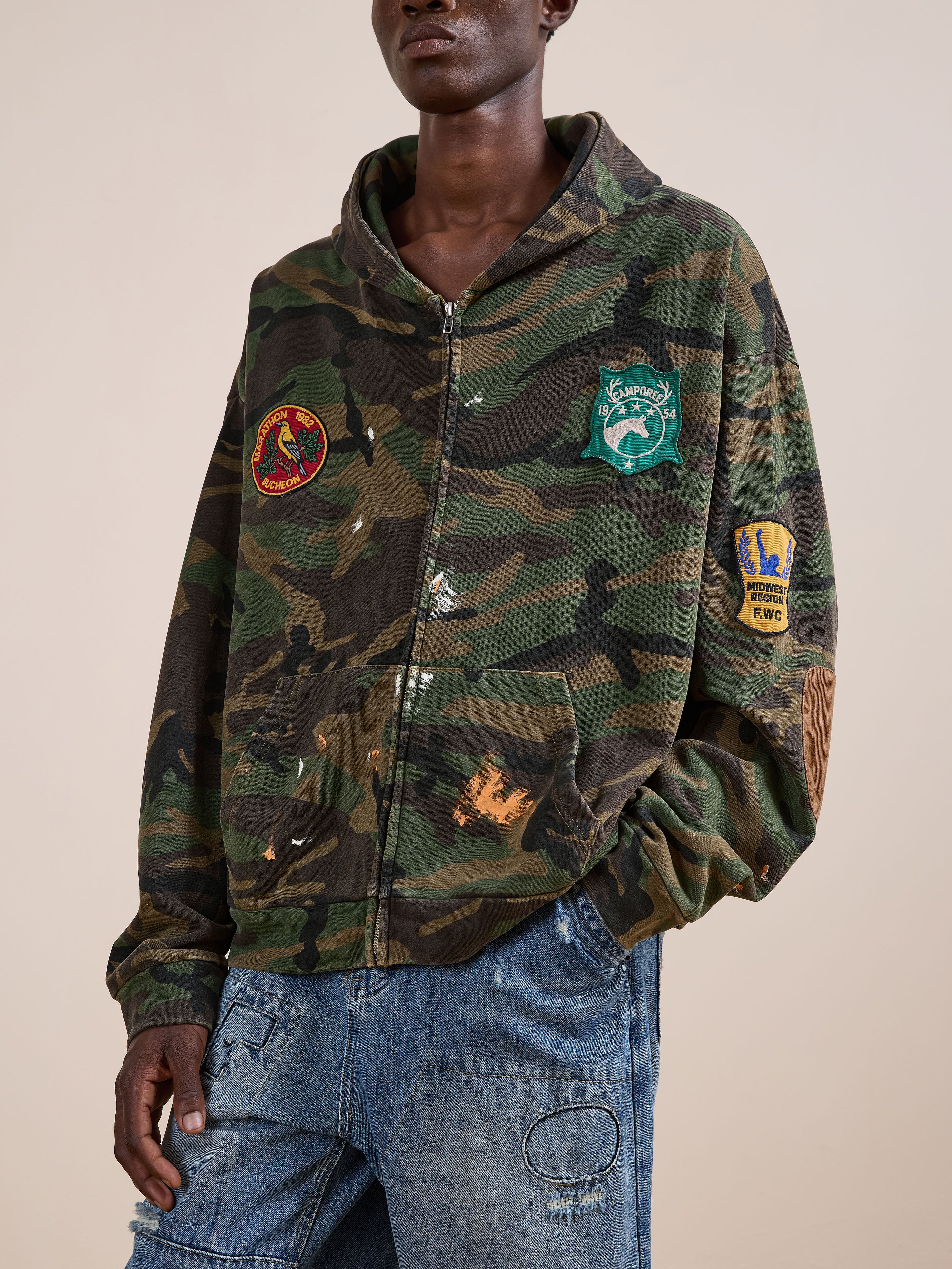 A person wears the FOUND Patch Camo Zip Up Hoodie, styled with relaxed fit blue jeans that have visible stitching and a knee patch.