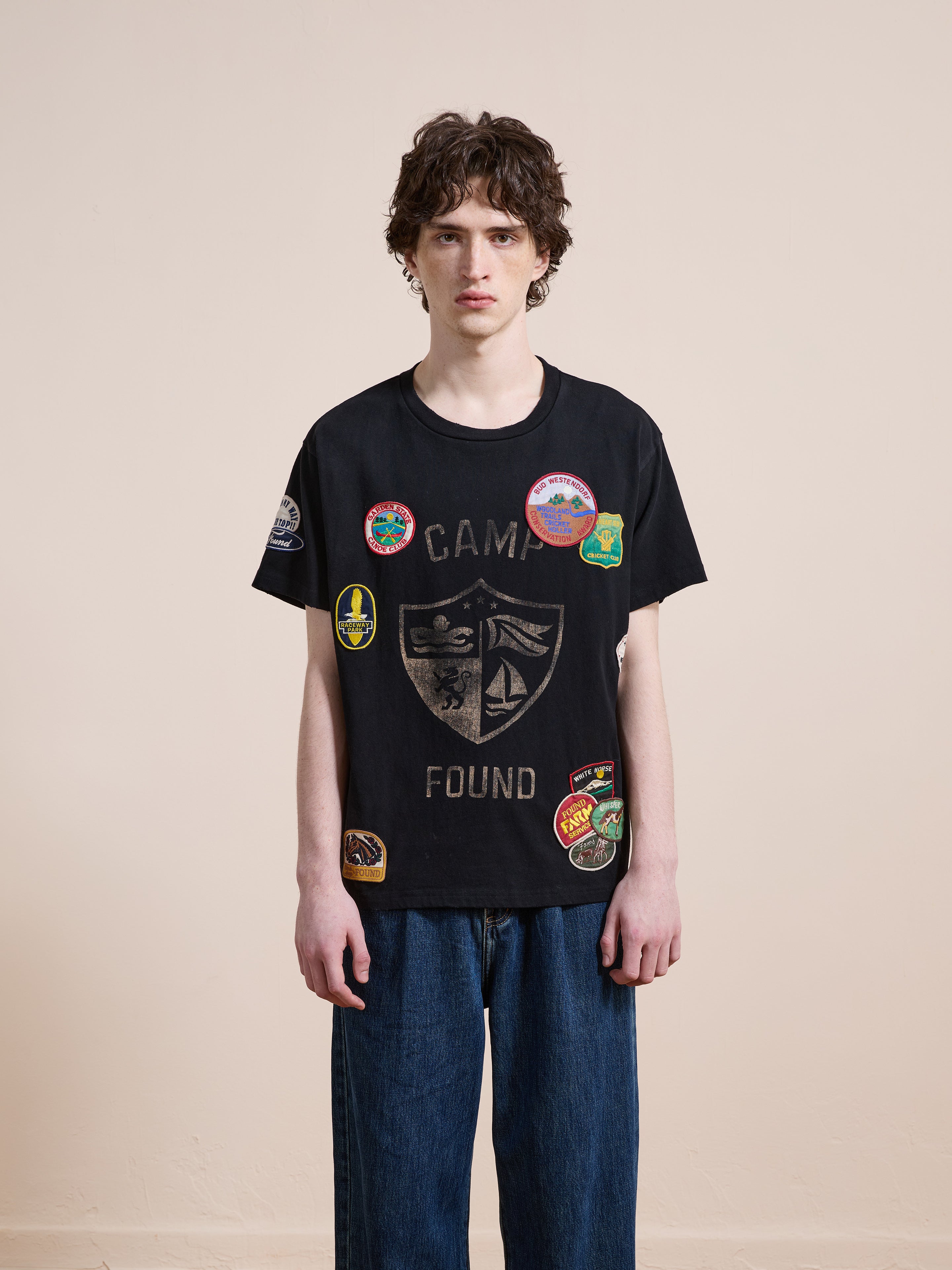A young person wears the FOUND Patchwork Camp Tee—a vintage black unisex shirt with "CAMP FOUND" text and colorful patches—styled with loose blue jeans for a relaxed, classic look against a plain background.