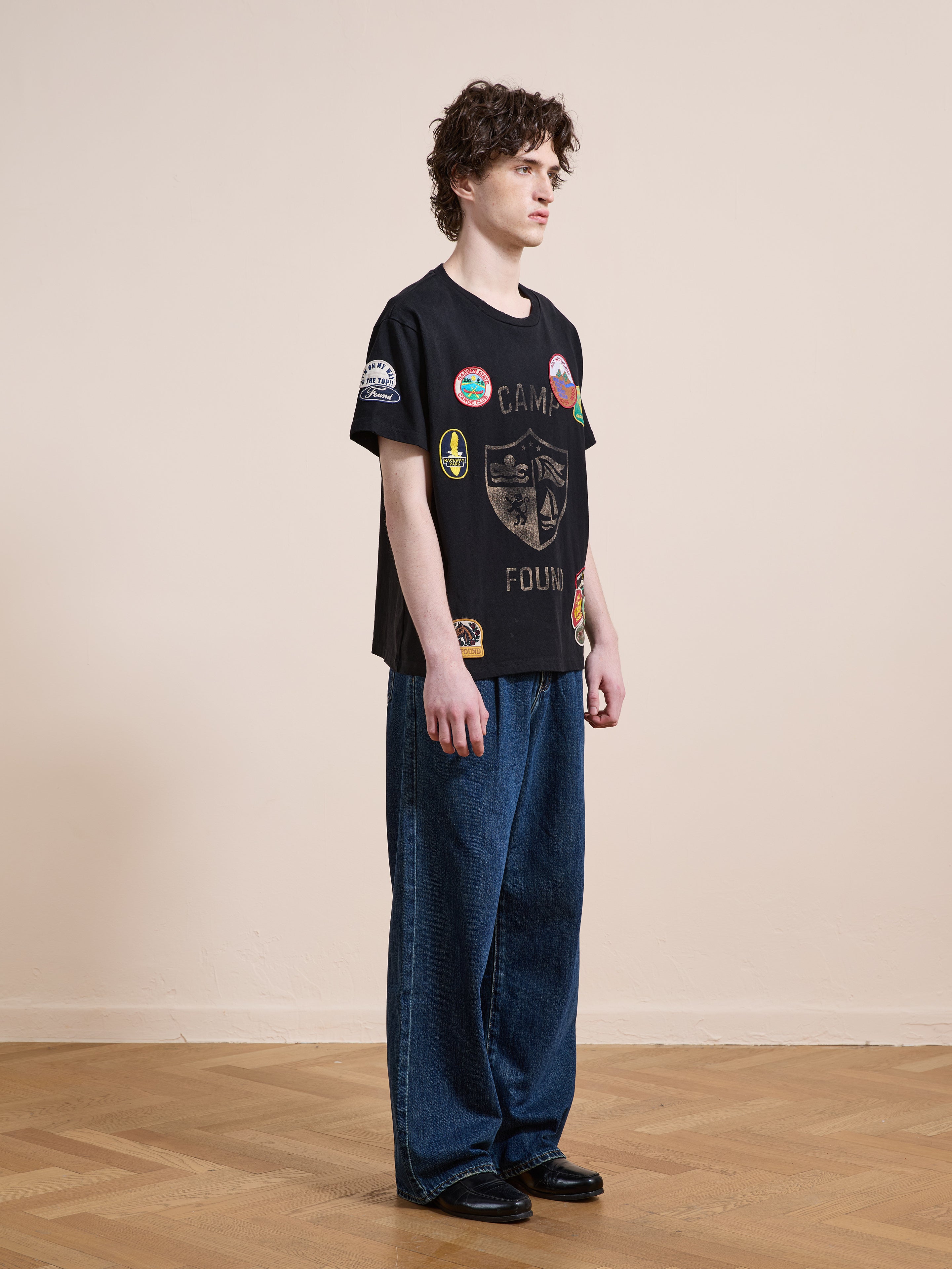 A person indoors wears the FOUND Patchwork Camp Tee, a unisex relaxed-fit vintage black shirt with patches, styled with wide-leg blue jeans and black shoes, standing sideways on a wooden floor against a beige wall.