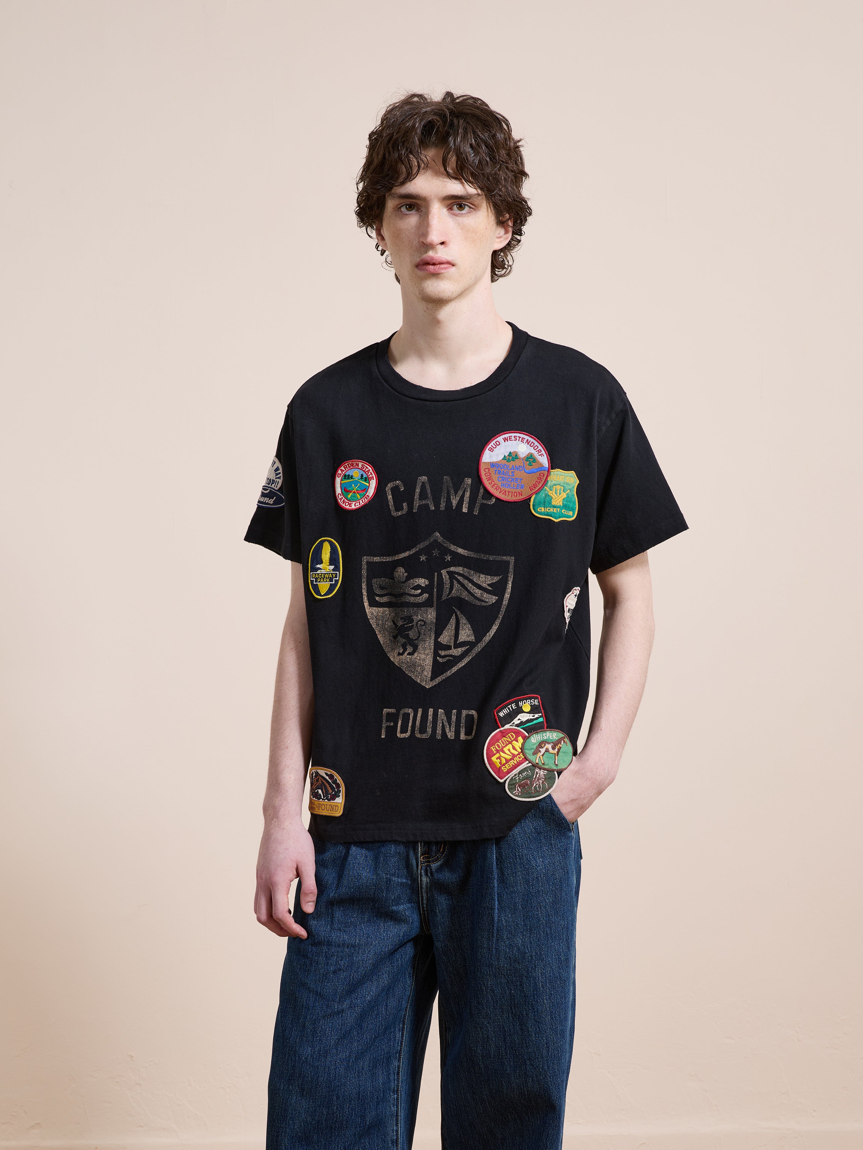 A young person with curly hair models the FOUND Patchwork Camp Tee, styled with blue jeans, against a plain beige background.