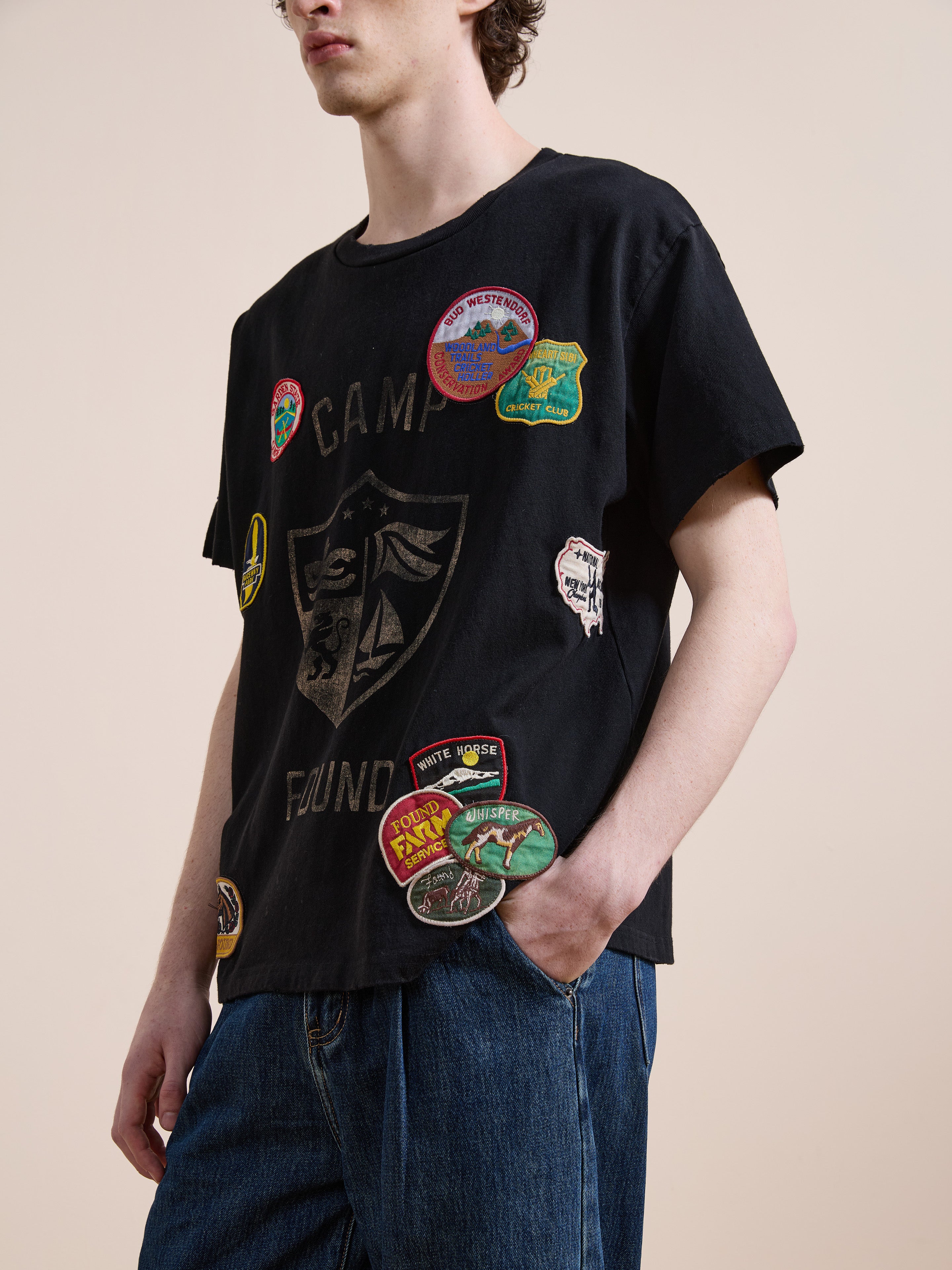 A person in the FOUND Patchwork Camp Tee—vintage black with colorful patches—and blue jeans stands with one hand in their pocket against a plain background.