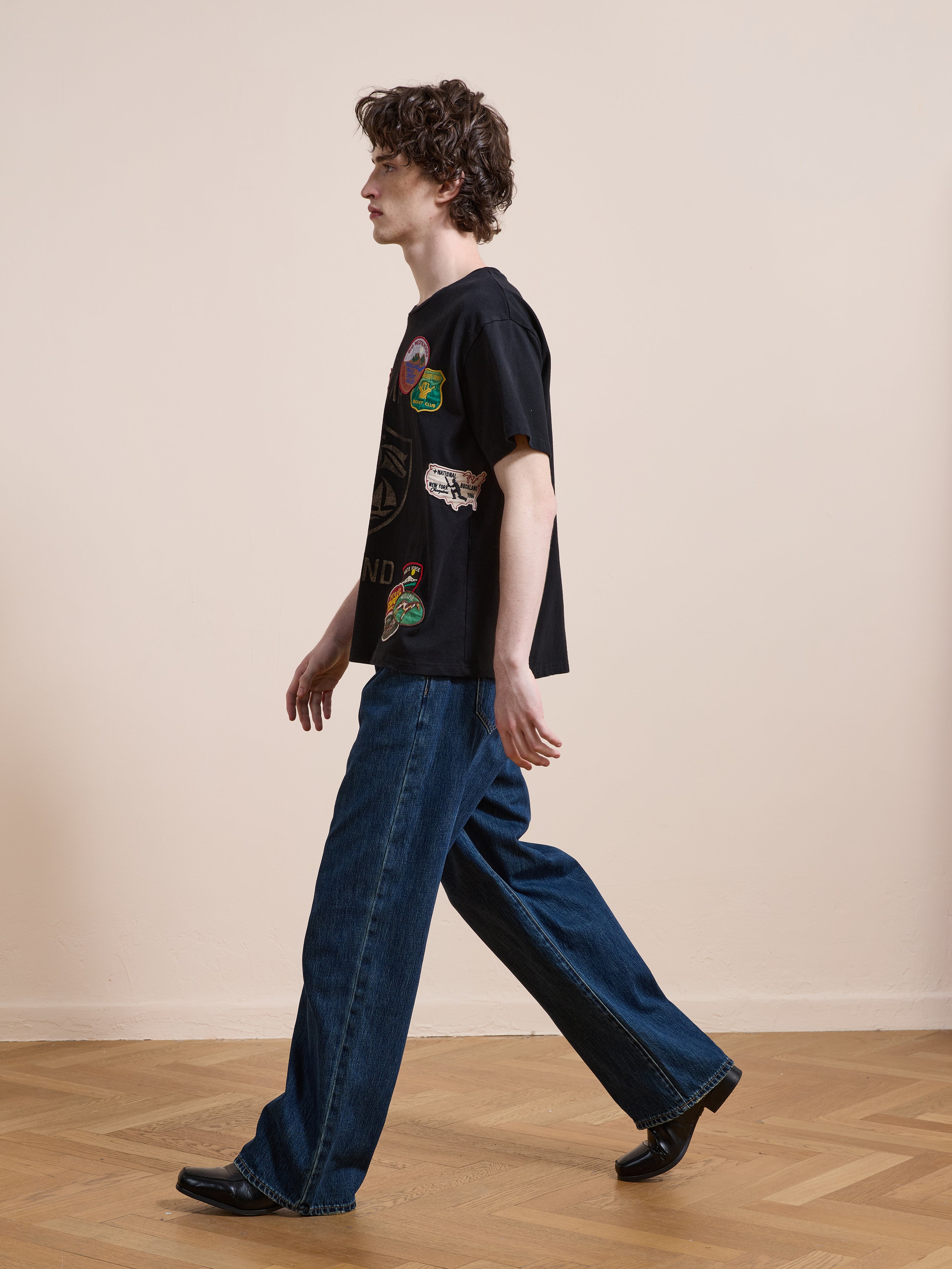 A person with curly hair walks indoors on a wooden floor, wearing the FOUND Patchwork Camp Tee in a unisex relaxed fit, paired with loose blue jeans and black shoes against a plain light-colored wall.
