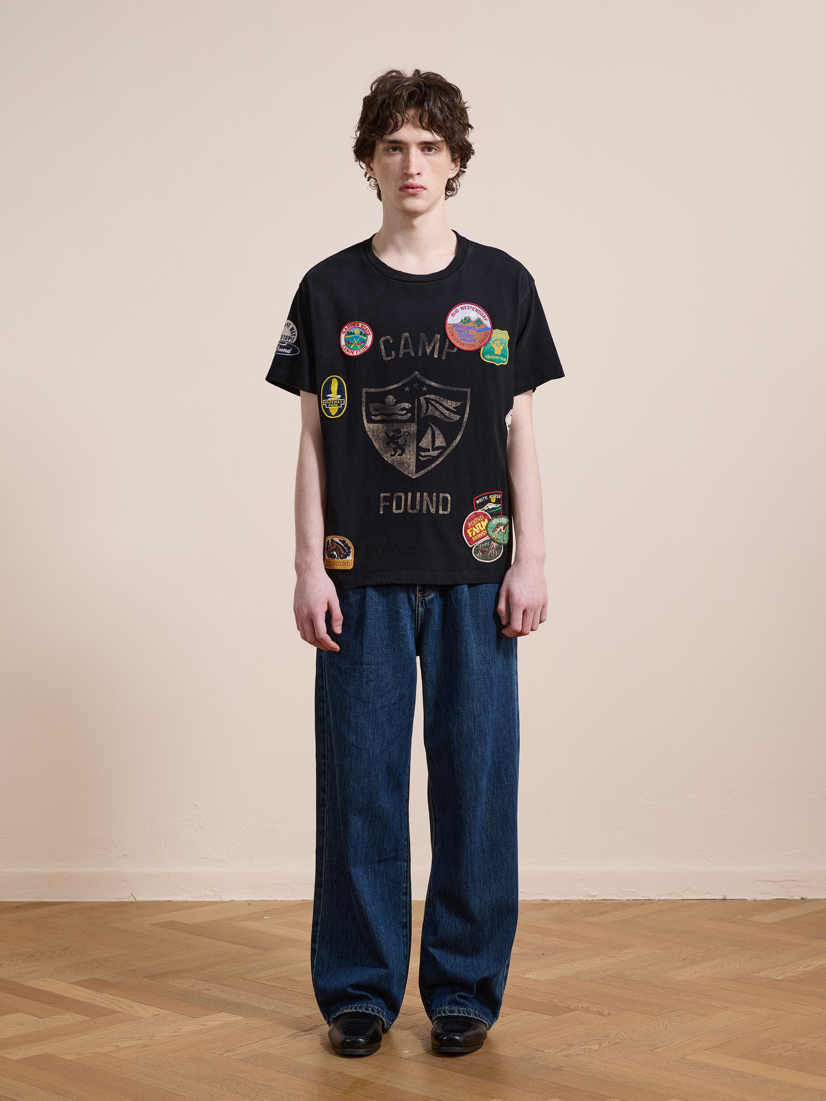 A person stands indoors against a plain wall, wearing the FOUND Patchwork Camp Tee and wide-leg blue jeans for a cool unisex relaxed look.