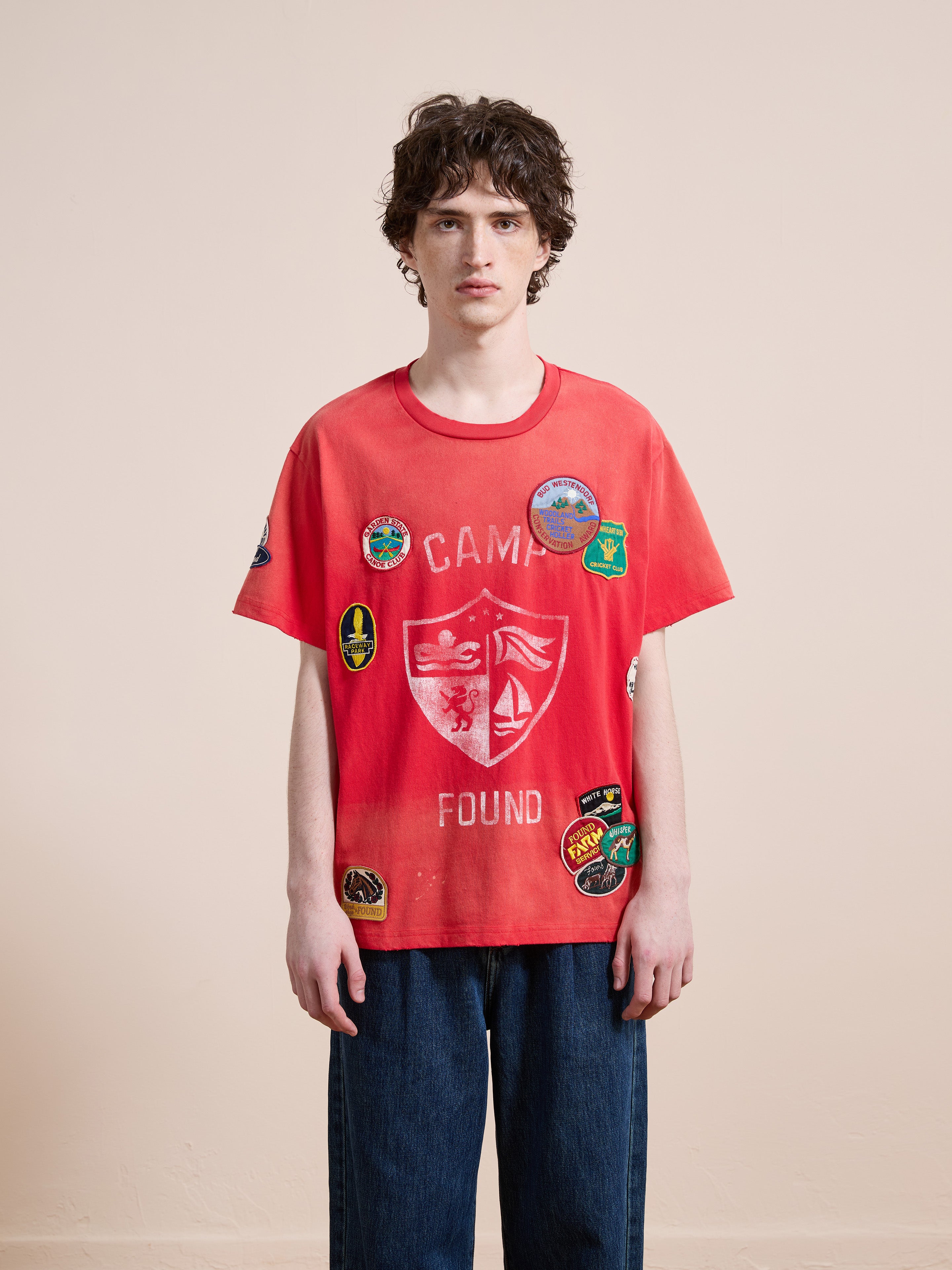 A young person wears the FOUND Patchwork Camp Tee in washed red—a unisex relaxed-fit cotton t-shirt with embroidered patches—paired with blue jeans against a plain background.