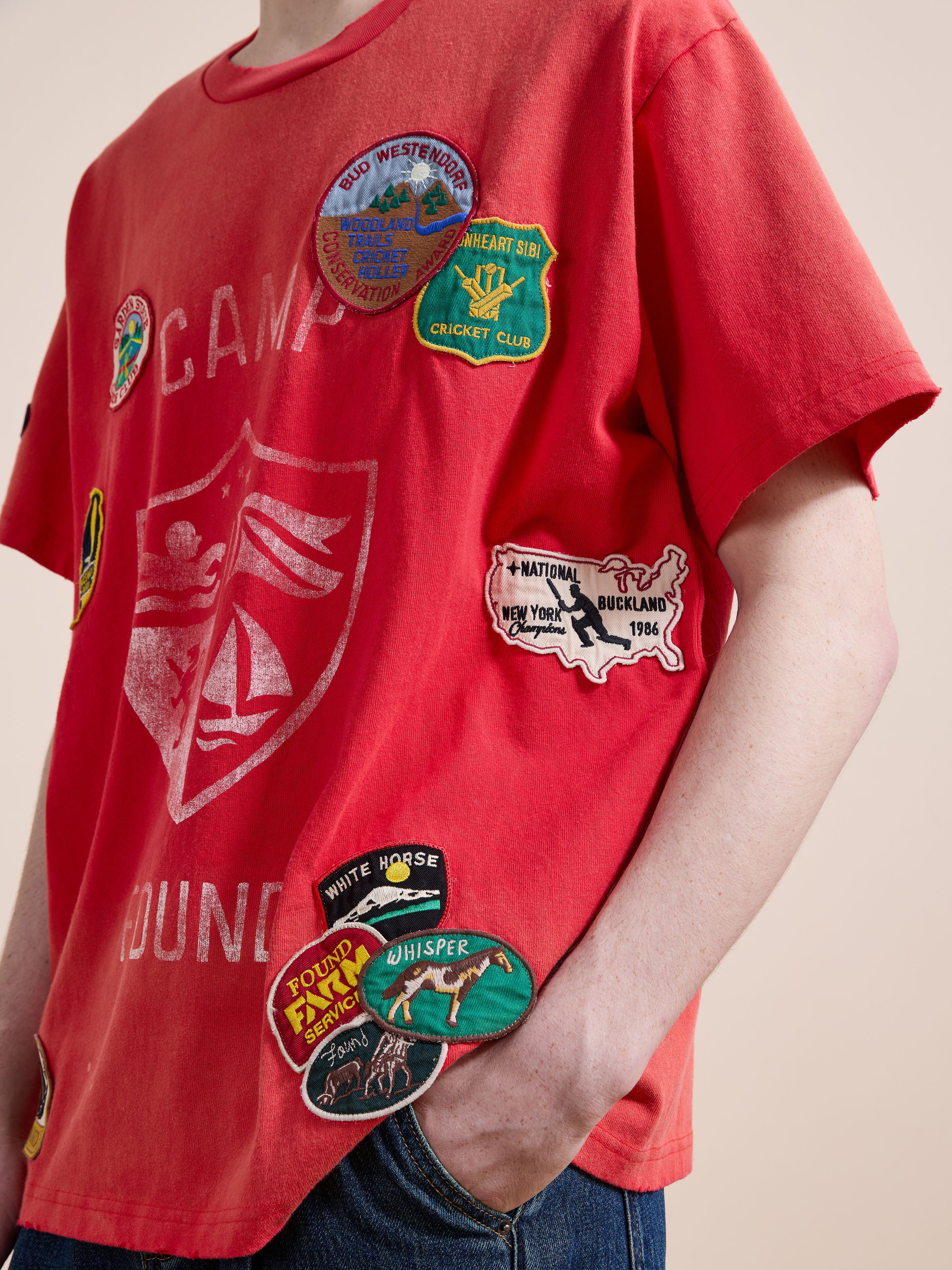 A person wears the FOUND Patchwork Camp Tee, a unisex cotton shirt featuring colorful embroidered patches, with one hand in their jeans pocket.