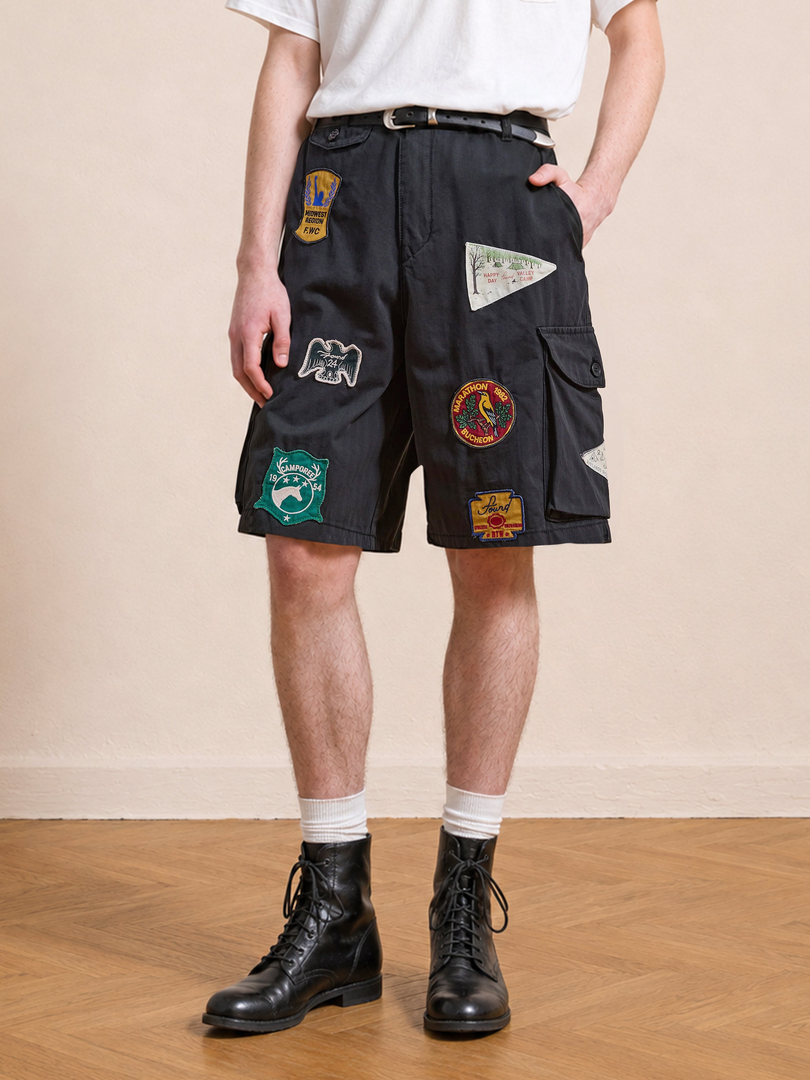Someone wearing Found Patchwork Cargo Shorts in vintage black with colorful patches, paired with a white t-shirt, white socks, and black lace-up boots, standing on a wooden floor.
