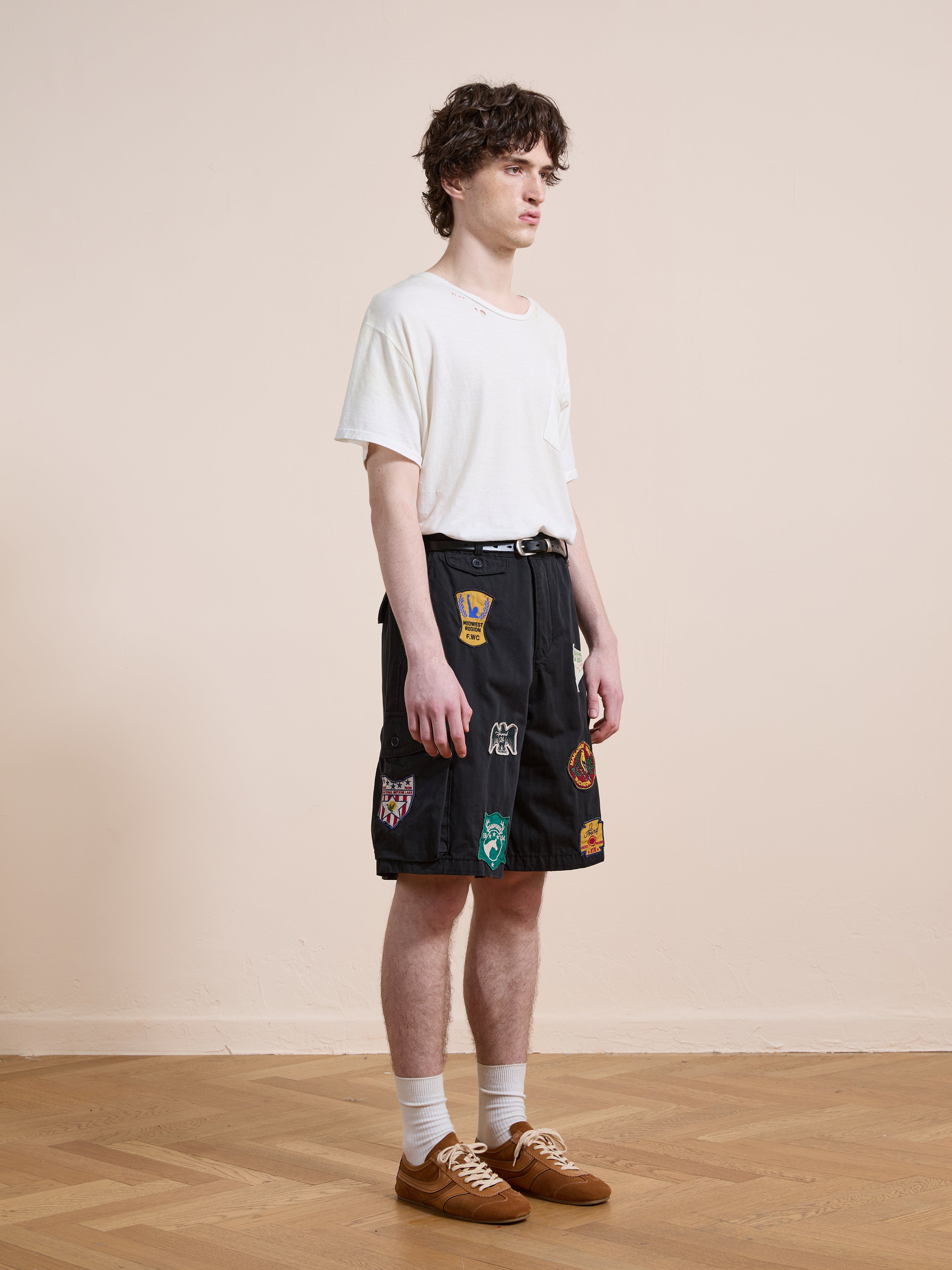 A young man stands indoors wearing a white t-shirt, Found Patchwork Cargo Shorts in vintage black wash (unisex relaxed fit), white socks, and brown sneakers against a plain beige wall and wooden floor.