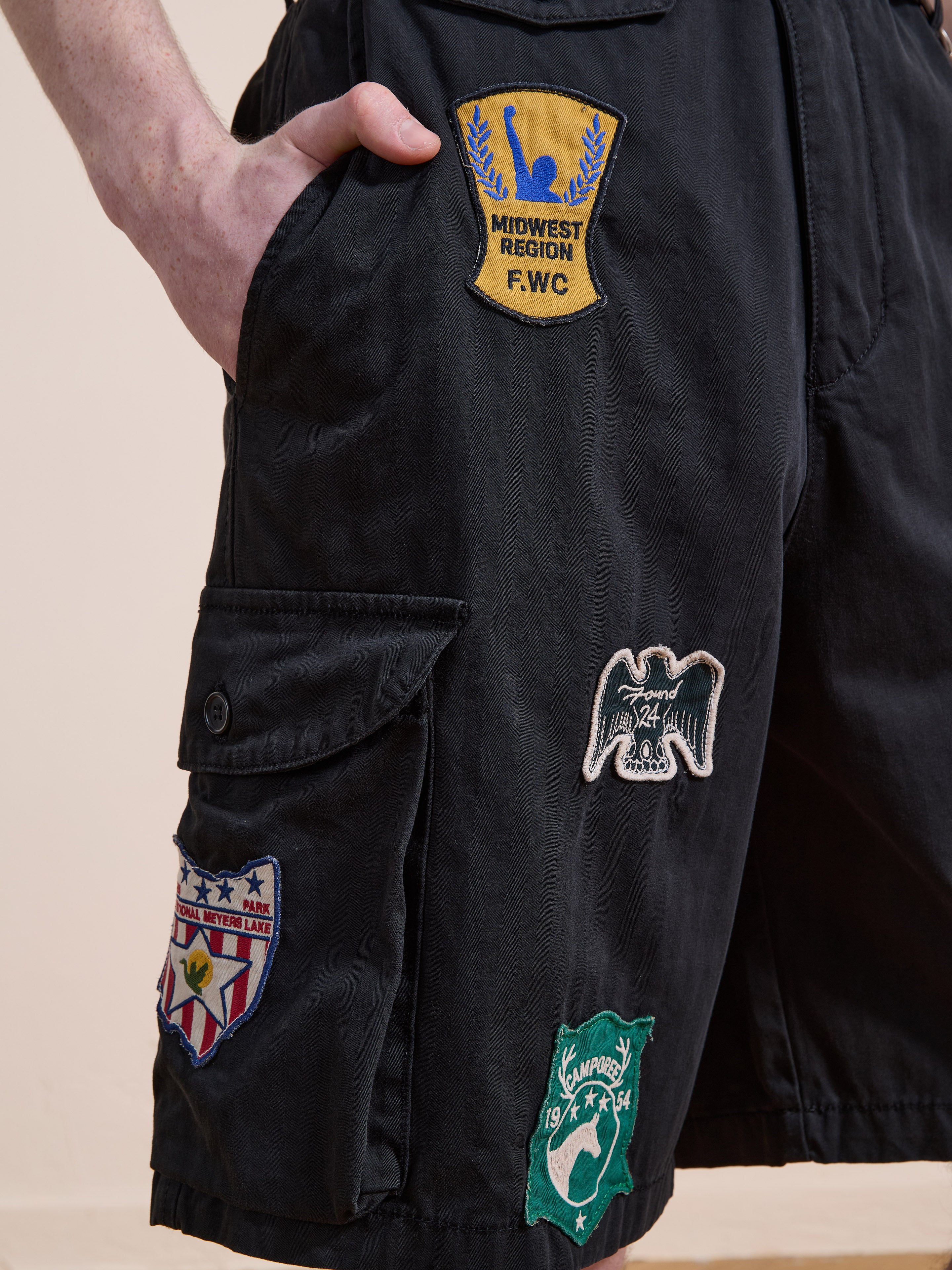 A person wears Found Patchwork Cargo Shorts, a unisex relaxed fit style in vintage black wash, detailed with embroidered logo patches on the front and side pocket.