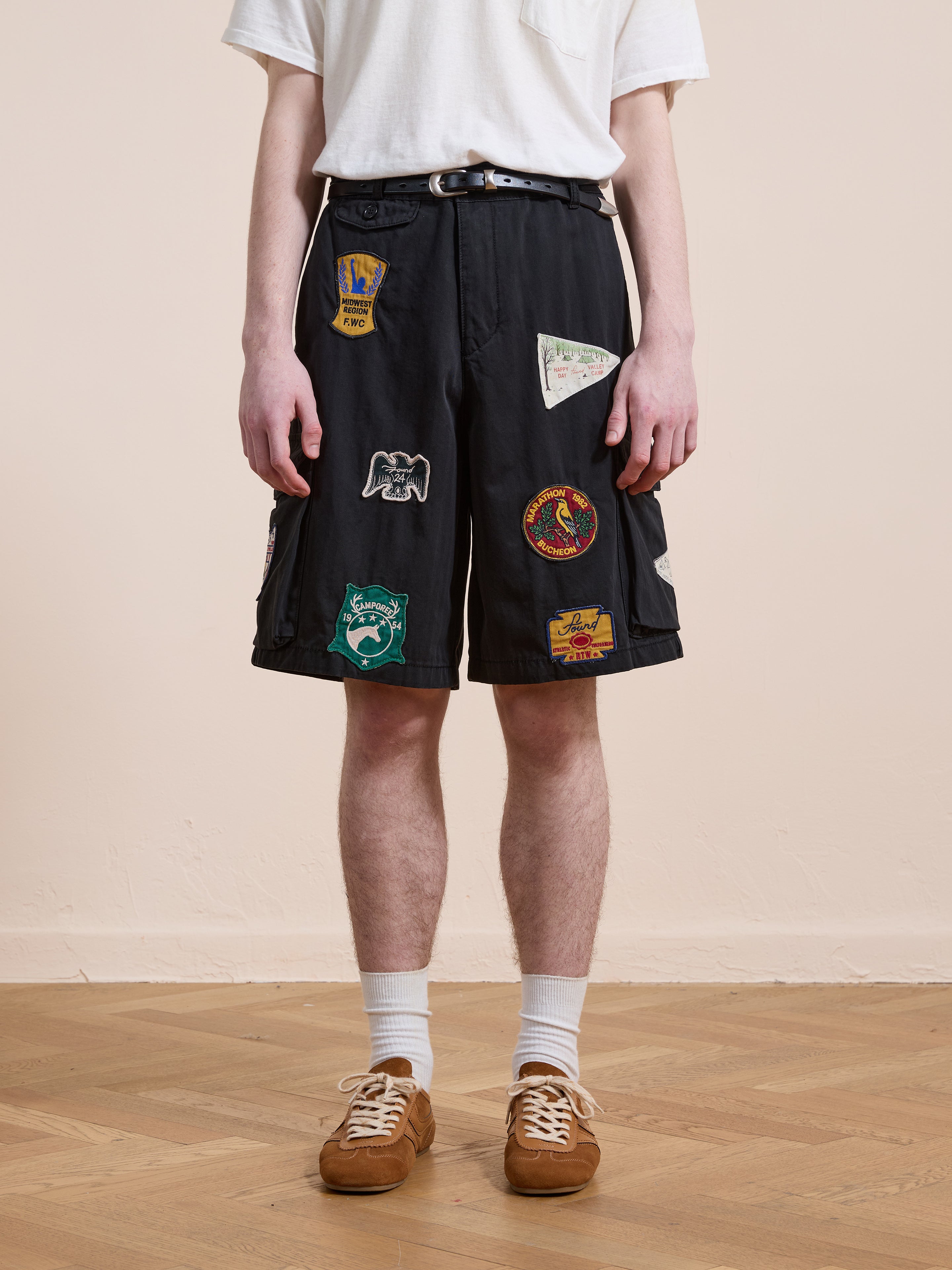 A person models the Found Patchwork Cargo Shorts—vintage black with colorful patches—paired with a white t-shirt, white socks, and brown sneakers on a wooden floor against a plain background.
