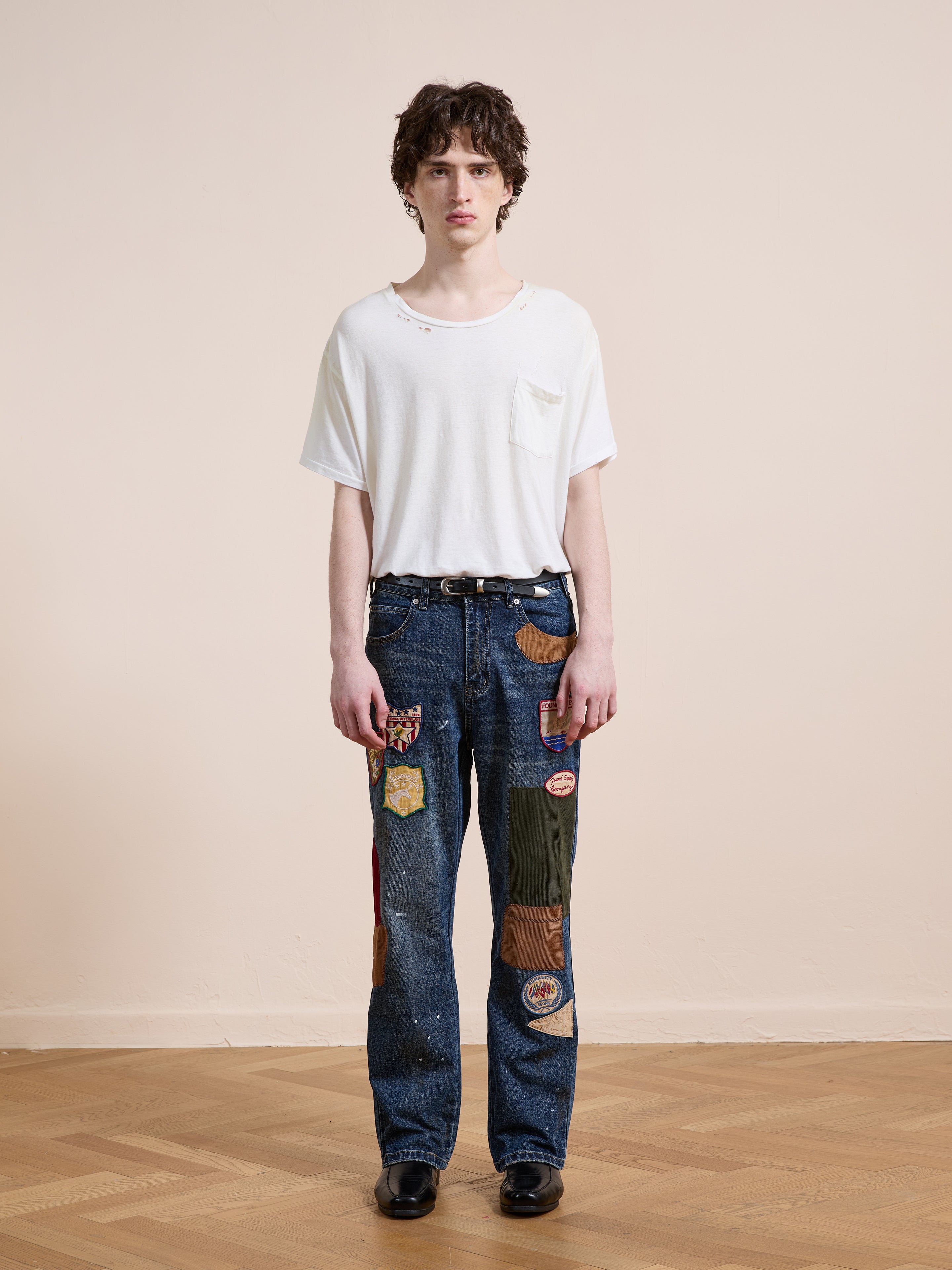 A person stands indoors on a wooden floor, wearing a white T-shirt and FOUND Patchwork Jeans with colorful patches, paired with black shoes.