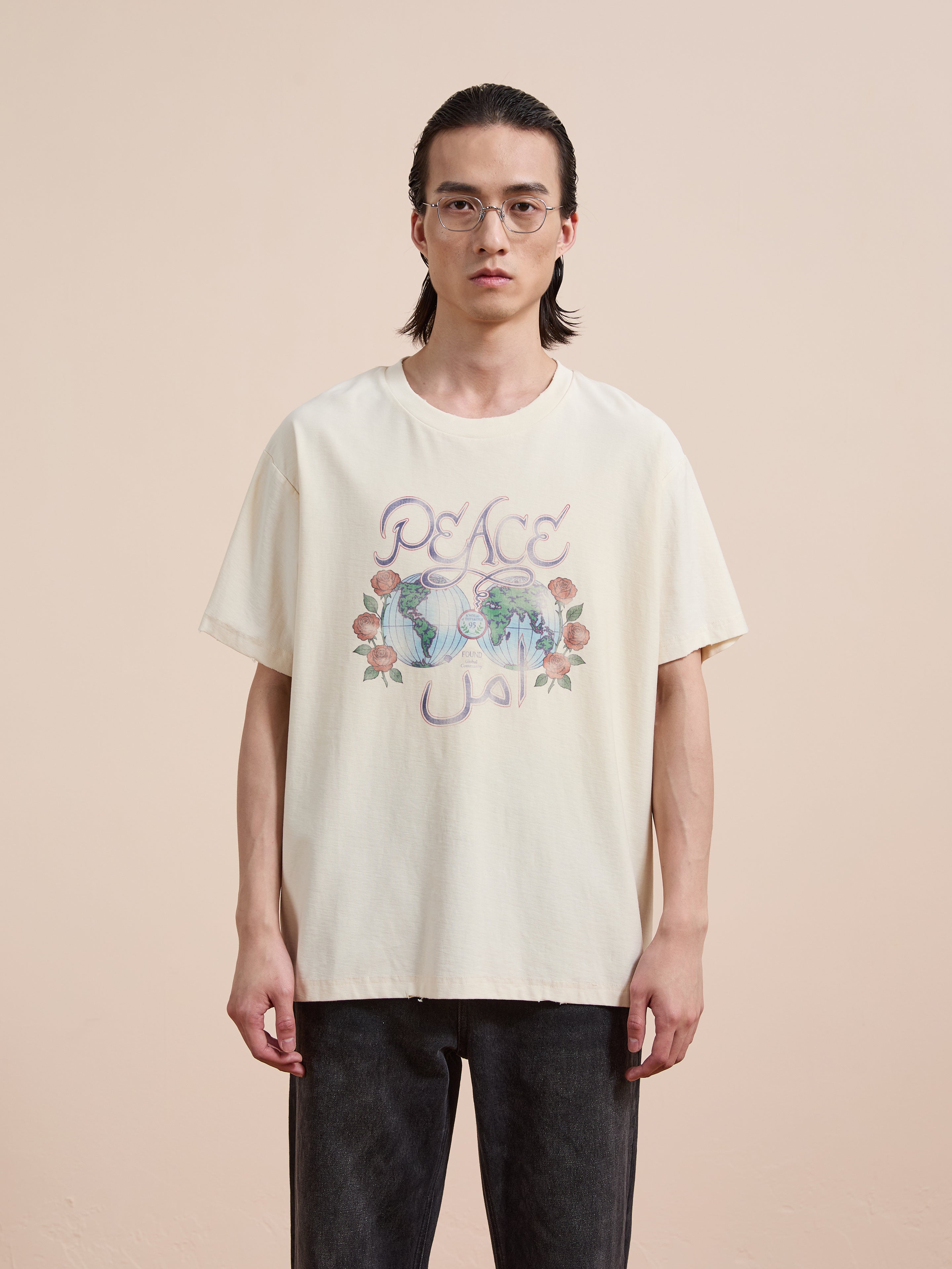 A person with long dark hair and glasses wears the FOUND Peace Globe Tee, a cream vintage cotton t-shirt with floral graphics and a unisex relaxed fit, standing against a plain beige background.