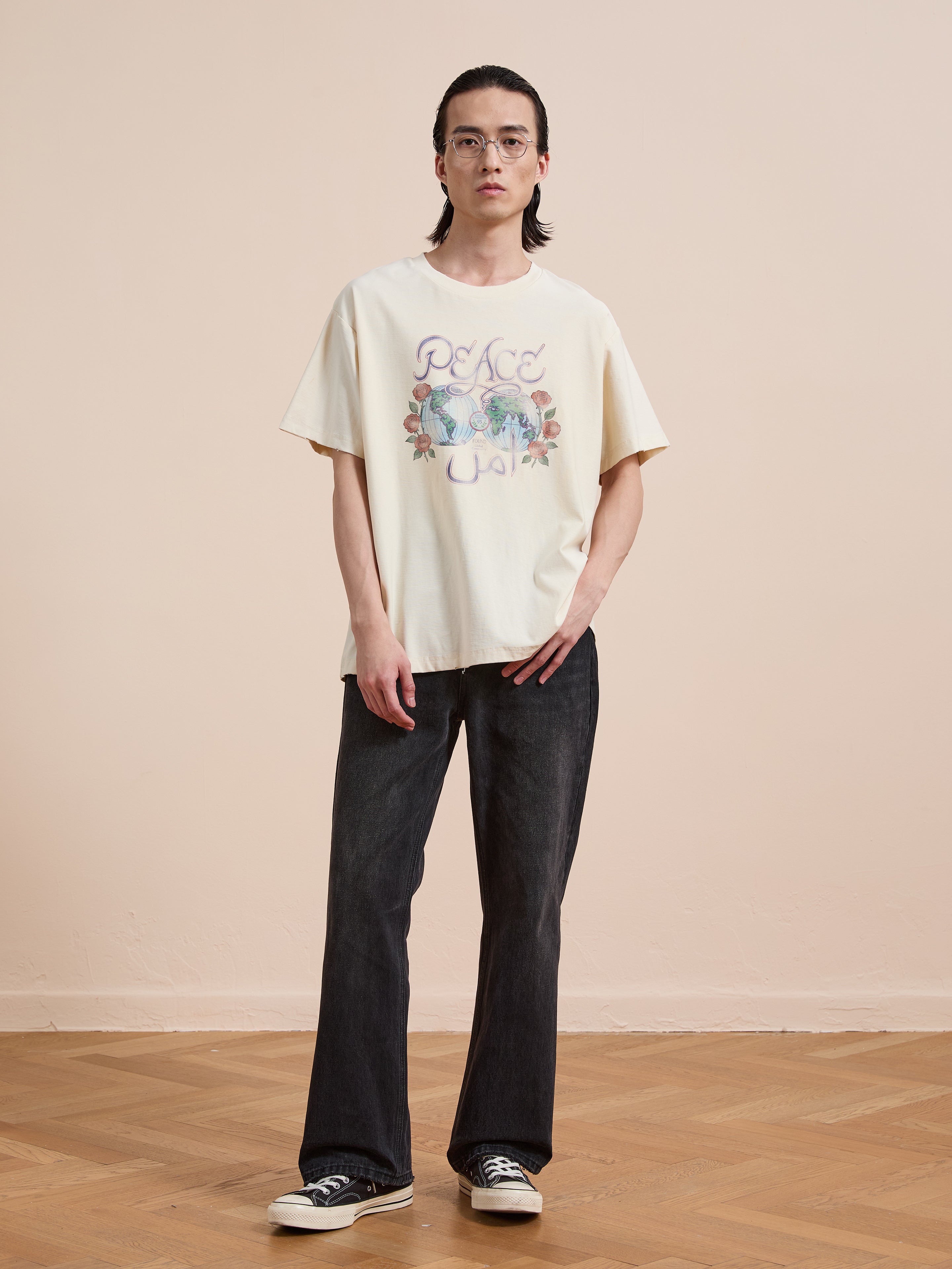 Person with long dark hair and glasses wears the FOUND Peace Globe Tee (unisex relaxed fit, cream), dark jeans, and black sneakers, standing on a wooden floor against a plain beige background.