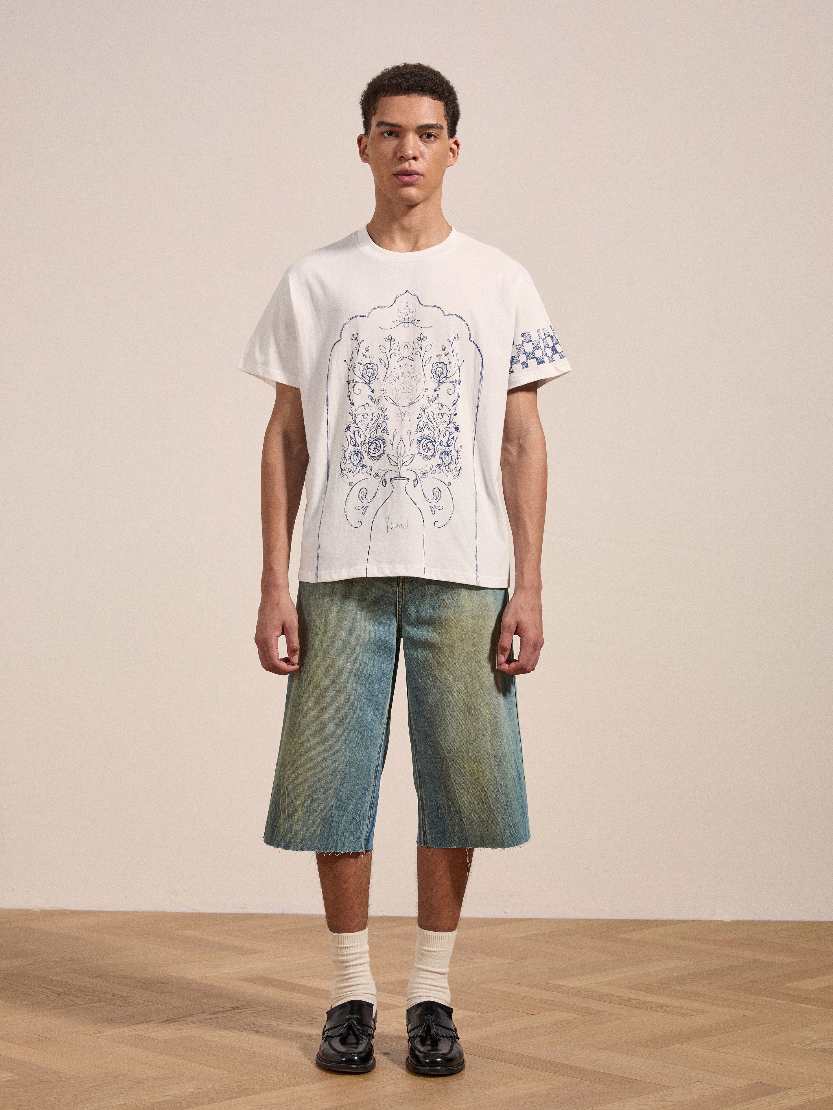 A man stands against a plain background wearing the "Pen Flower Medley" graphic t-shirt by FOUND, knee-length denim shorts, white socks, and black shoes.