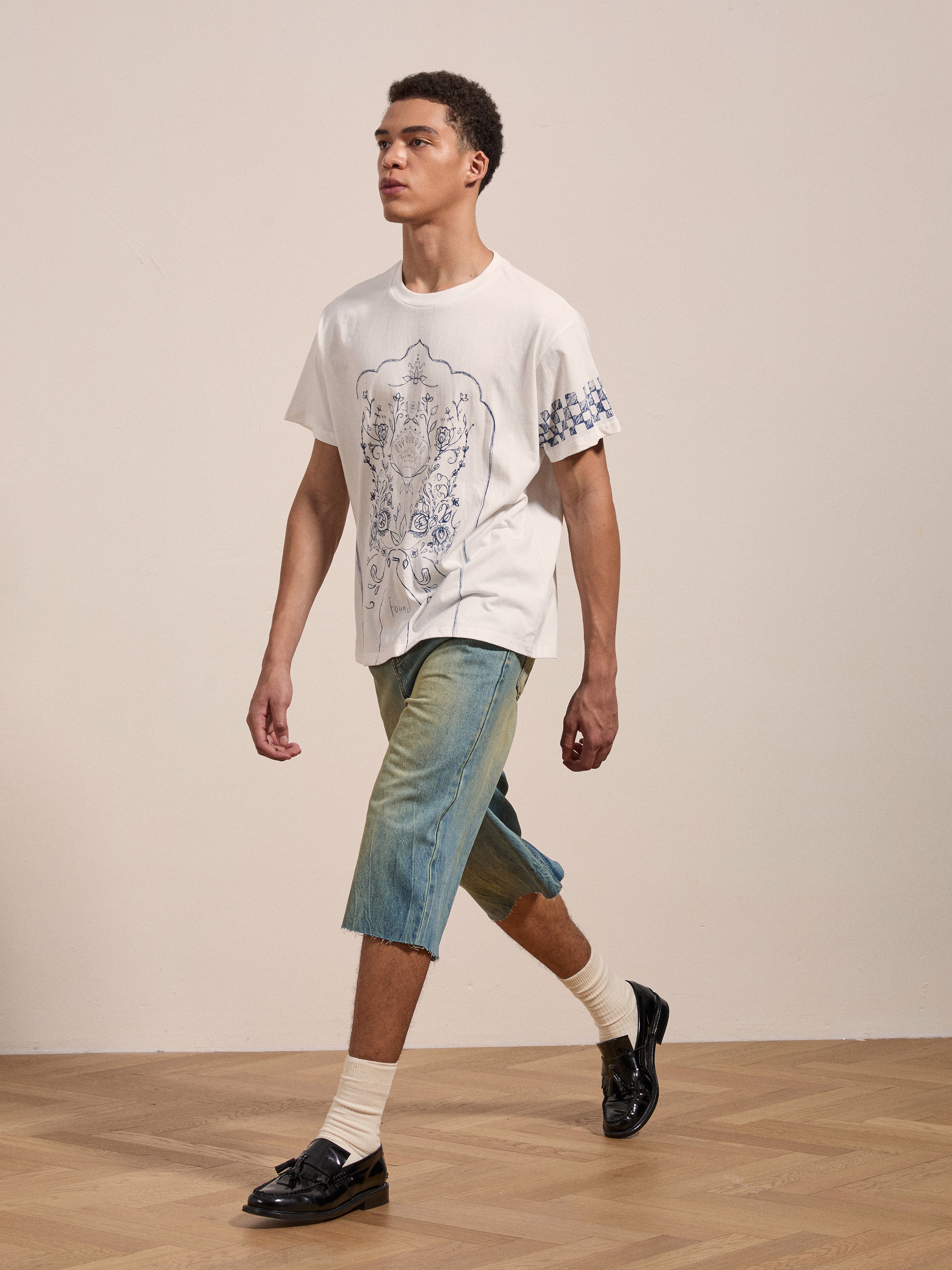 A man wearing a white FOUND "Pen Flower Medley" graphic t-shirt, denim knee-length shorts, white socks, and black loafers walks on a wooden floor against a plain background.