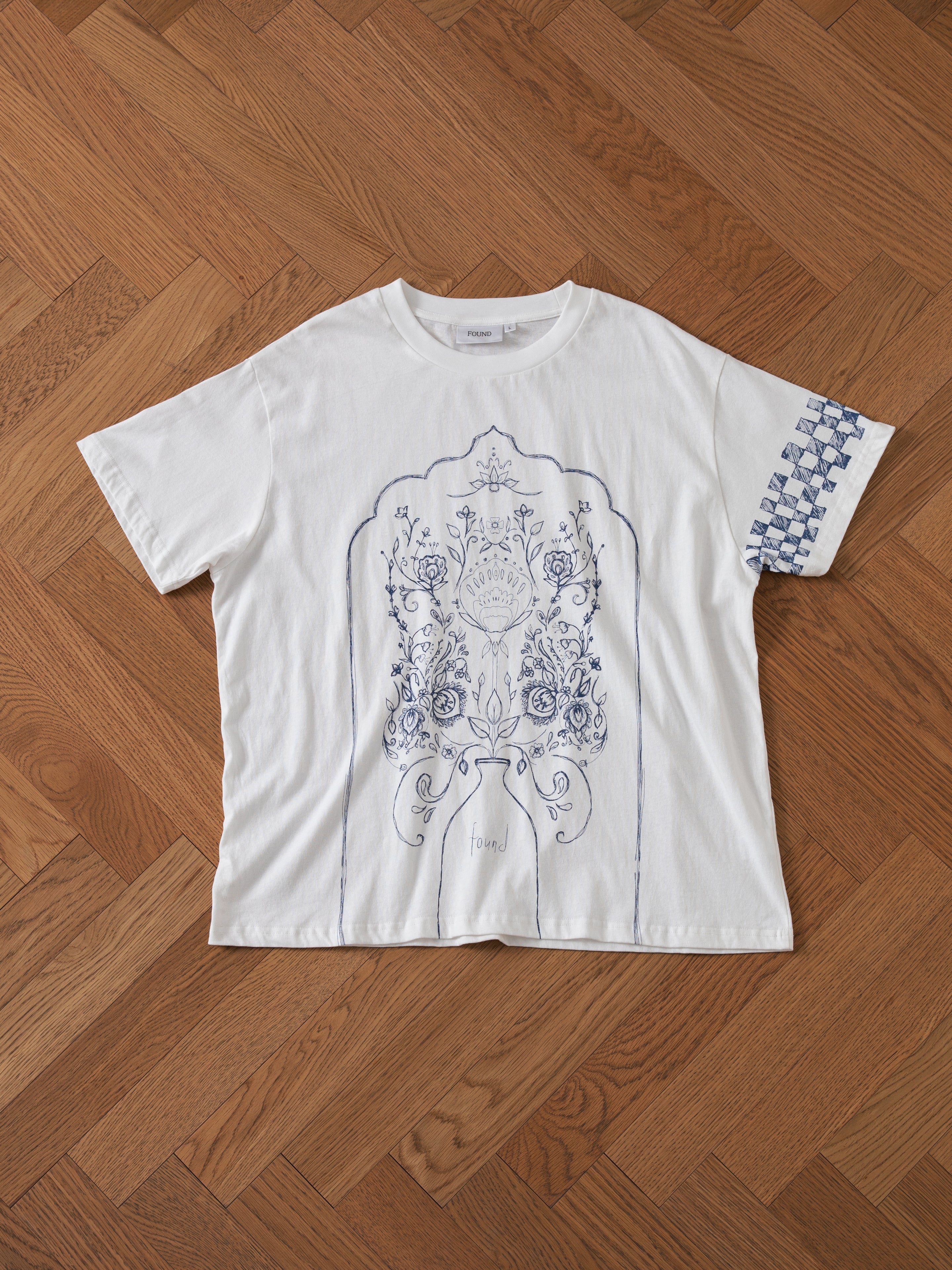The FOUND Pen Flower Medley T-shirt, featuring an intricate blue floral and geometric design on the front and sleeve, lies flat on a wooden floor with a herringbone pattern.