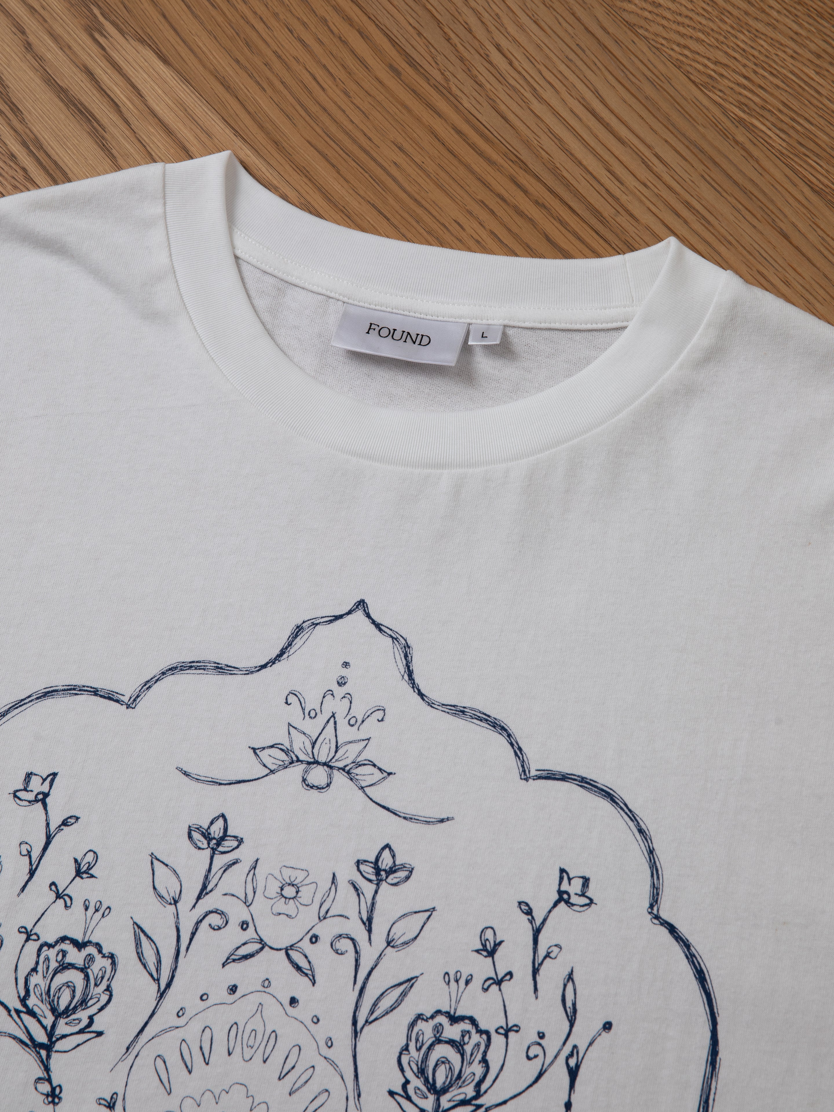 A close-up of the Pen Flower Medley white t-shirt, featuring a blue floral and pattern design on the front and a label that reads "FOUND" inside the neckline, laid on a wooden surface.