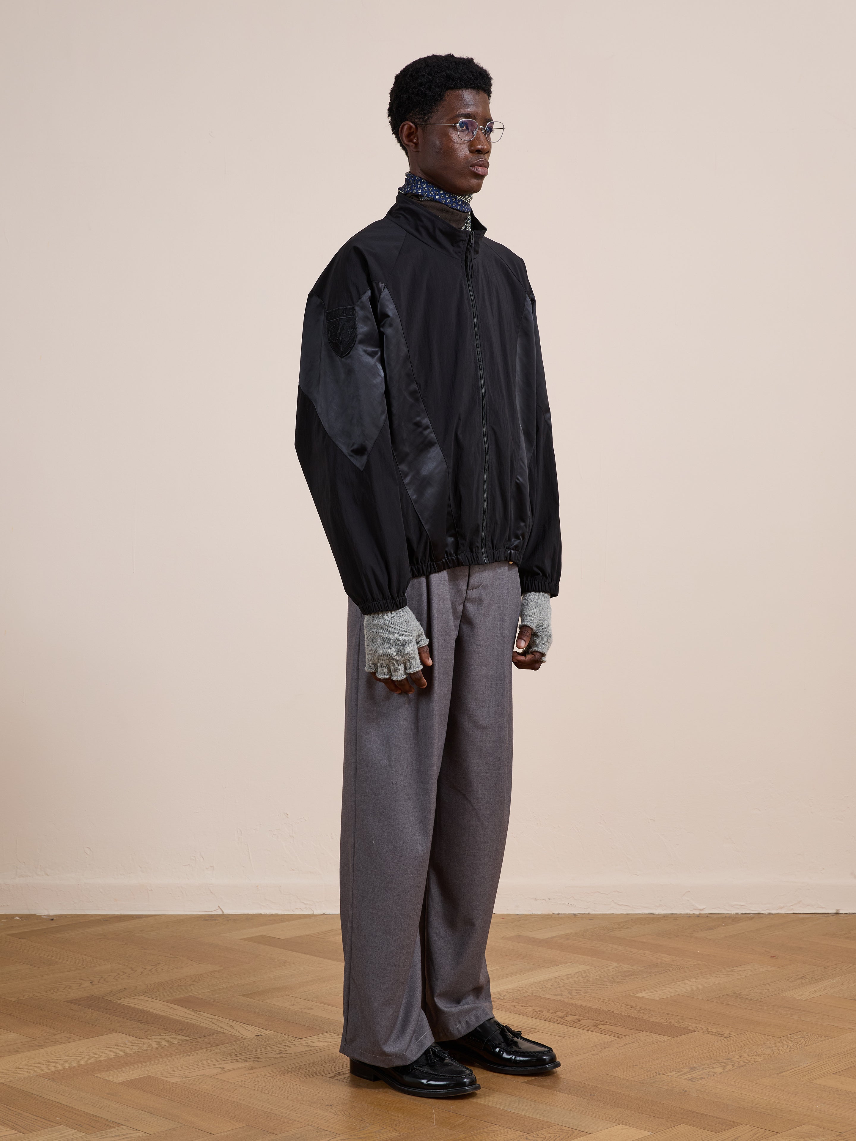 Indoors against a light wall, a person wears FOUND's Pine and Rose Jacket in black, paired with gray wide-leg pants, gray fingerless gloves, black shoes, and glasses.