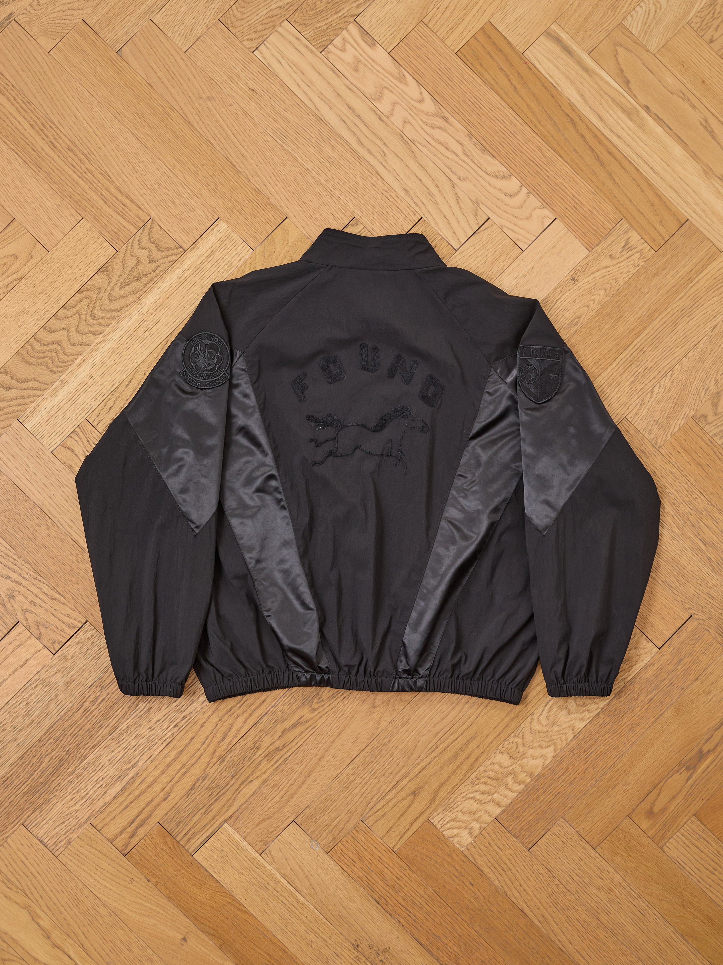 The Pine and Rose Jacket by FOUND is a black unisex windbreaker with "FOUND" and a running dog on the back, shiny sleeve panels, and circular upper-arm patches, adding vintage sportswear flair. Shown laid flat on wooden flooring.