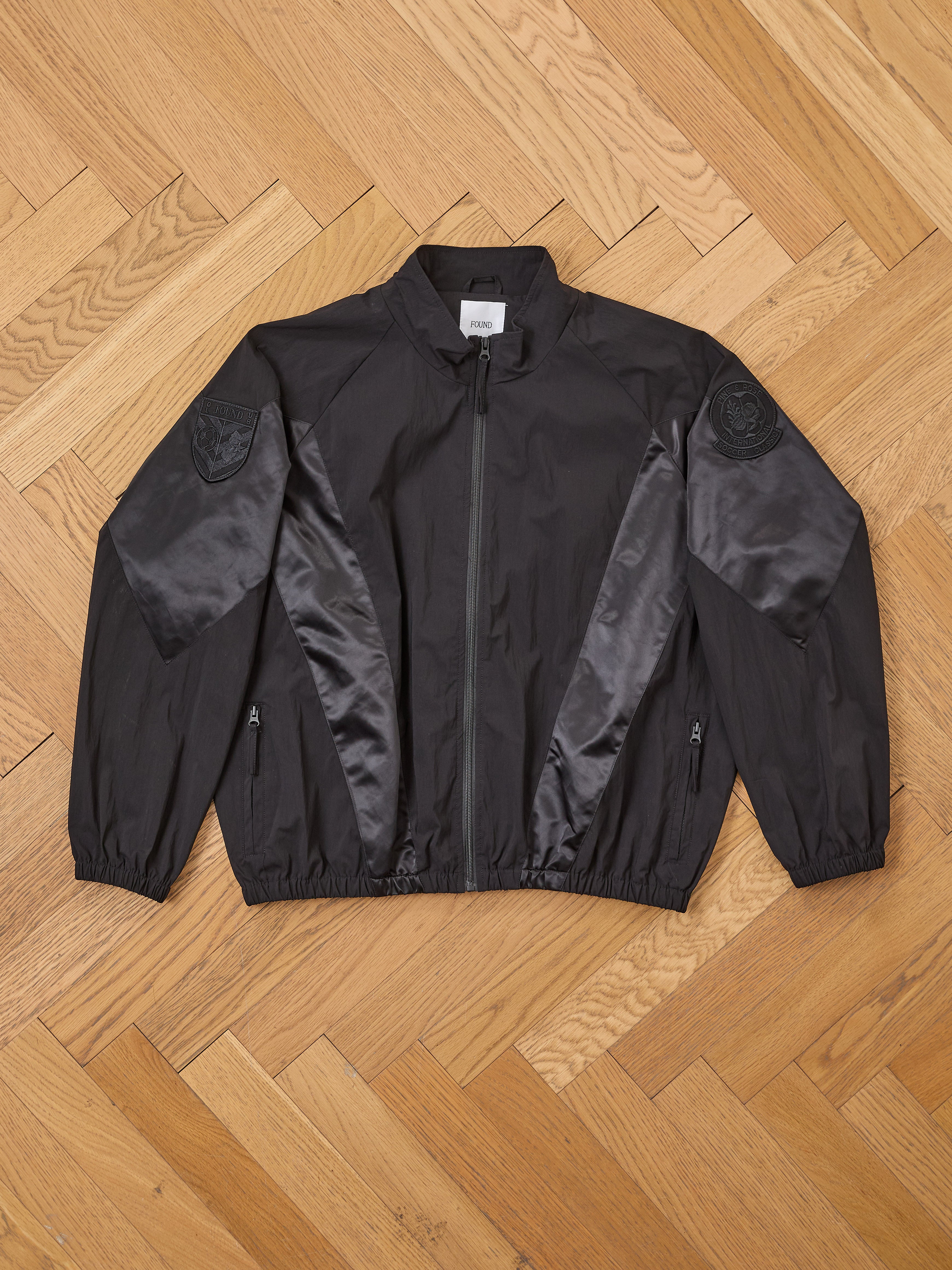 The FOUND Pine and Rose Jacket, a vintage sportswear-inspired black zip-up with satin panels and embroidered shoulder patches, is displayed flat on a light wooden herringbone floor.