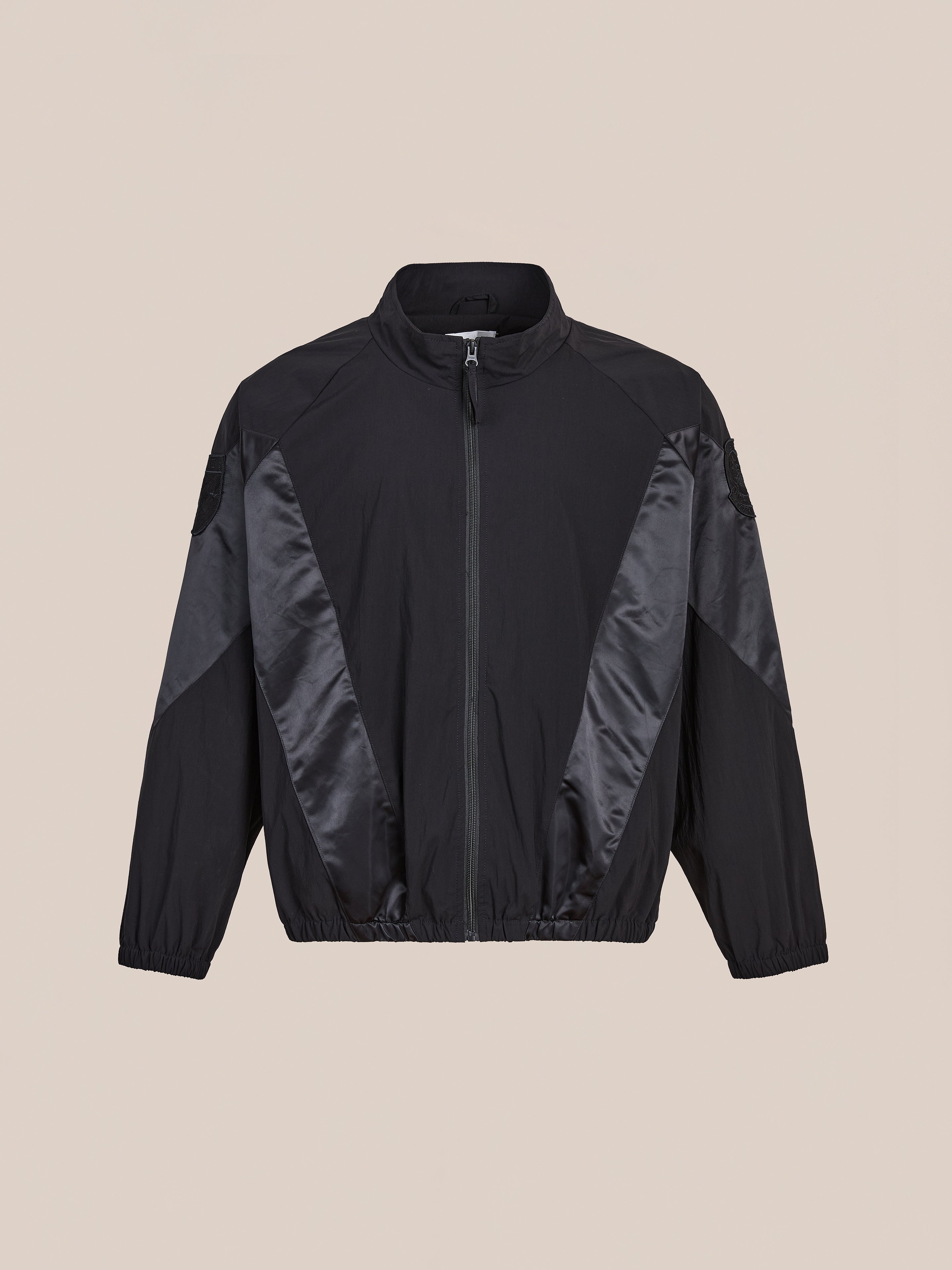 The FOUND Pine and Rose Jacket features a black zip-up design with a high collar, subtle sheen panels on the front and sleeves, elastic cuffs and waistband, showcased against a plain beige background for vintage sportswear appeal.