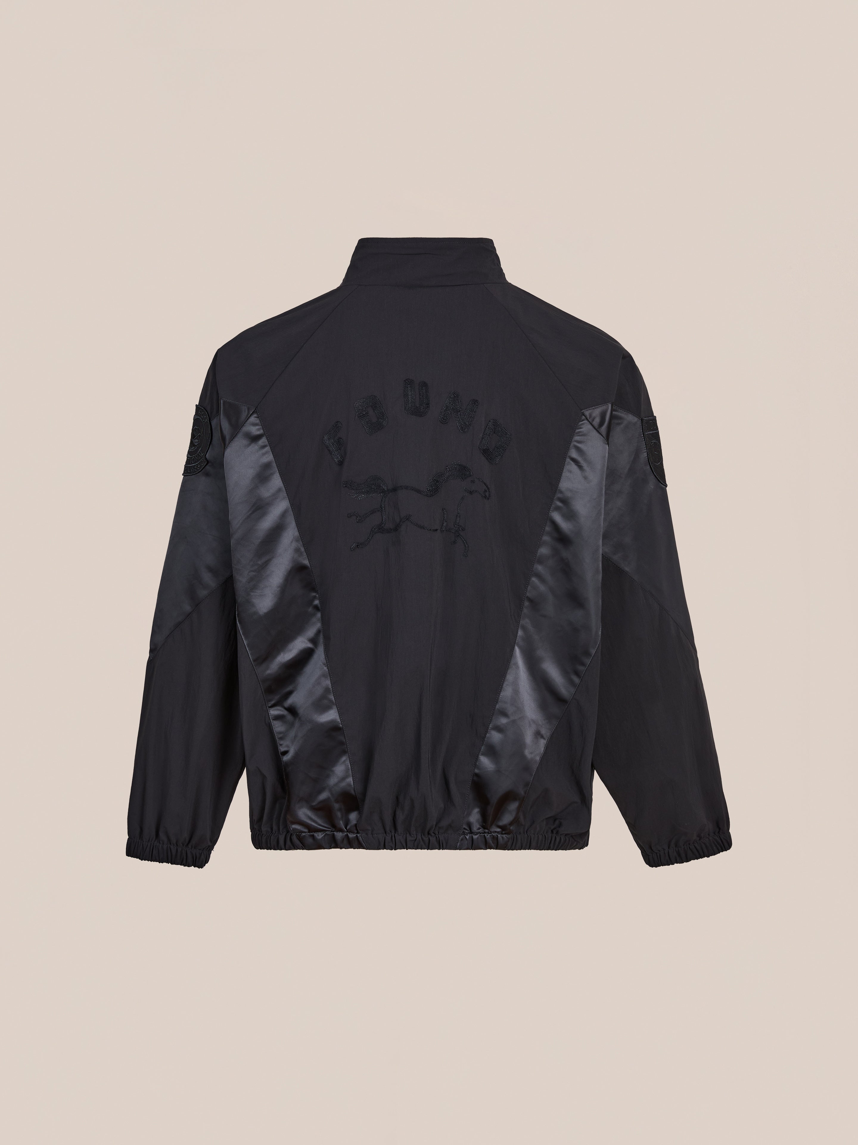 The Pine and Rose Jacket by FOUND is a black unisex zip-up windbreaker featuring shiny black side panels, elastic cuffs and hem, plus an embroidered horse graphic with "FOUND" on the back—a vintage-inspired sportswear look.