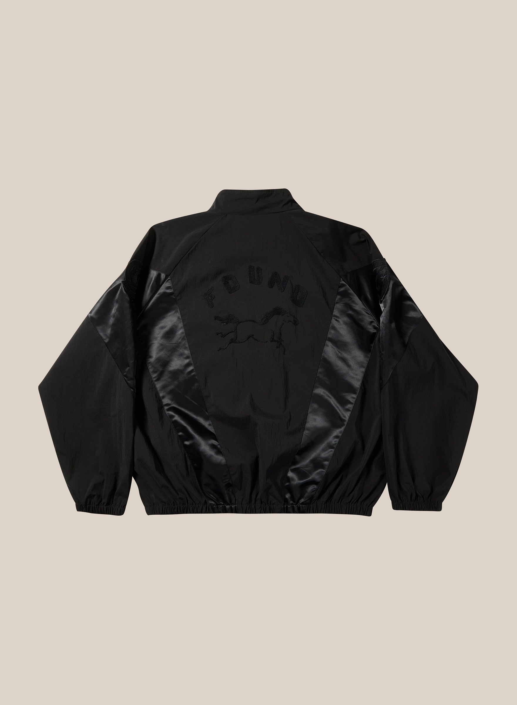 The Pine and Rose Jacket by FOUND is a black unisex windbreaker with shiny triangular panels on the back, an outlined horse design, and "FOUND" above it. Features include a collar, elastic cuffs, and hem for a vintage sportswear feel.