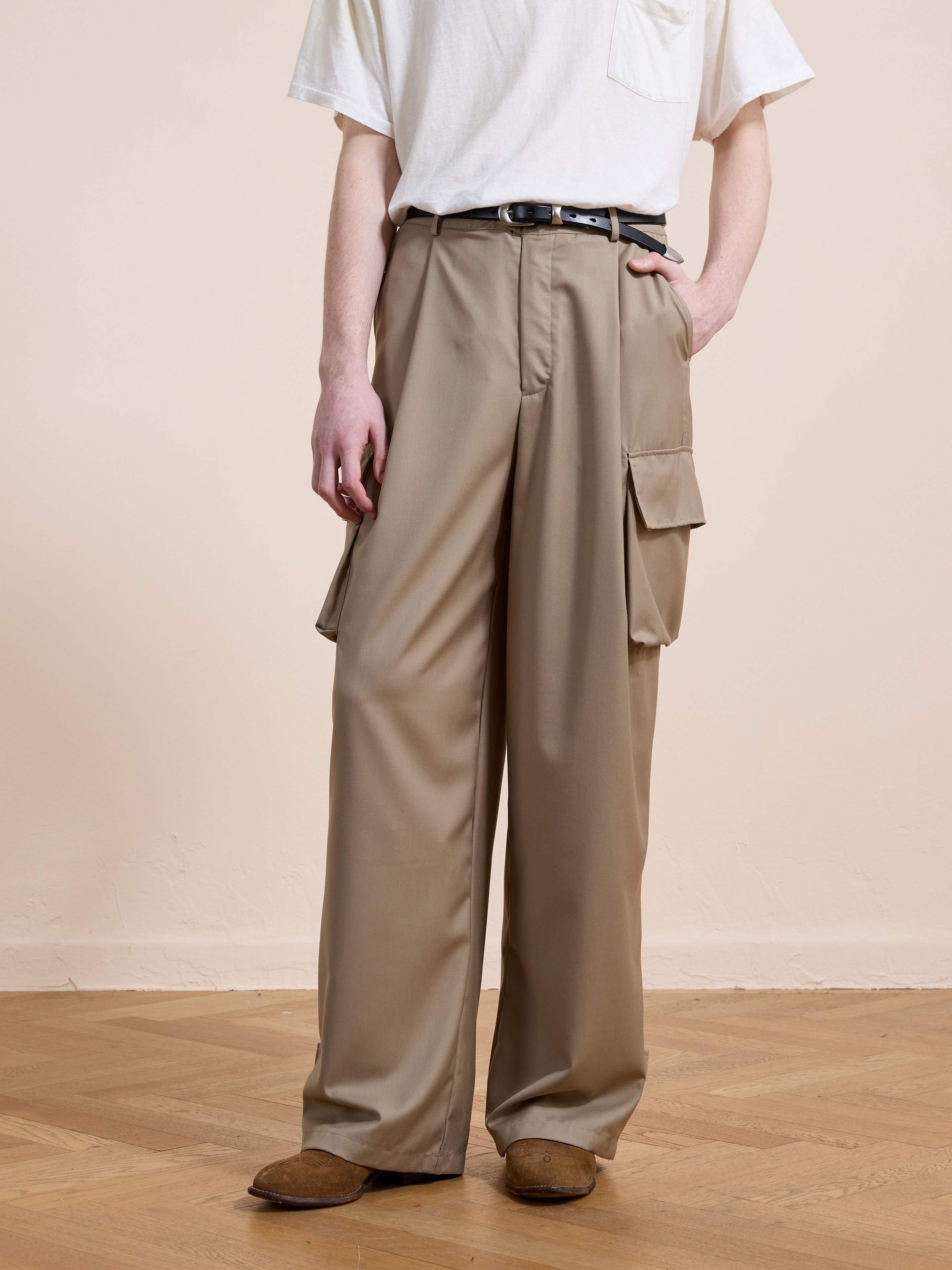 A person wears FOUND Pleated Cargo Pants—beige, relaxed fit with side pockets—paired with a white t-shirt, black belt, and brown shoes, standing on a wooden floor against a plain background.