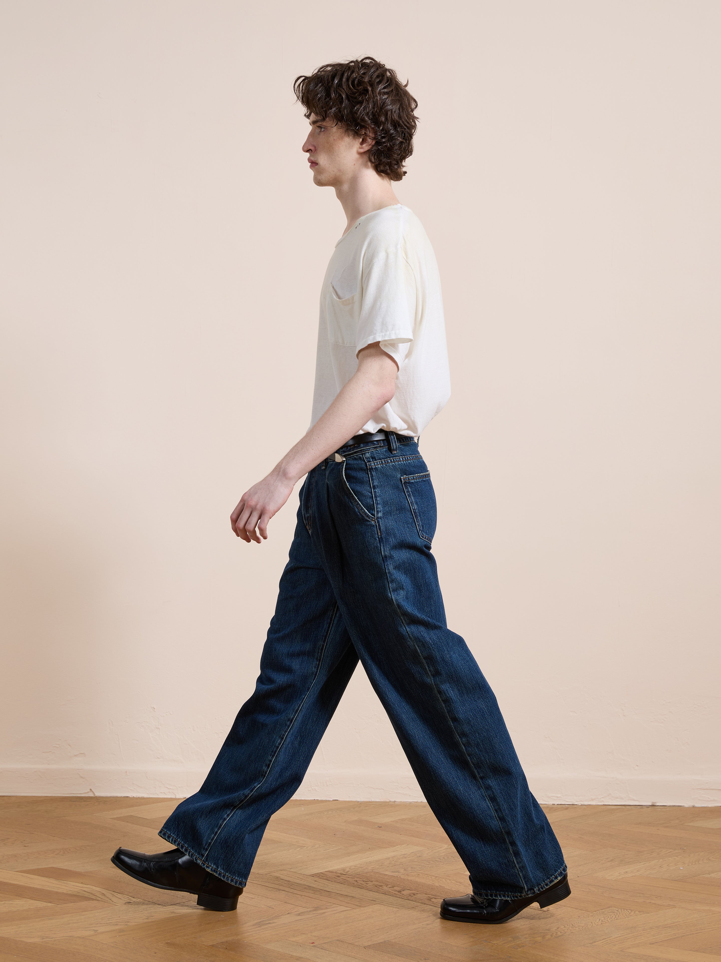 A person with curly hair walks indoors against a light wall and wooden floor, wearing Found Pleated Relaxed Fit Jeans in blue, a white t-shirt, and black shoes.