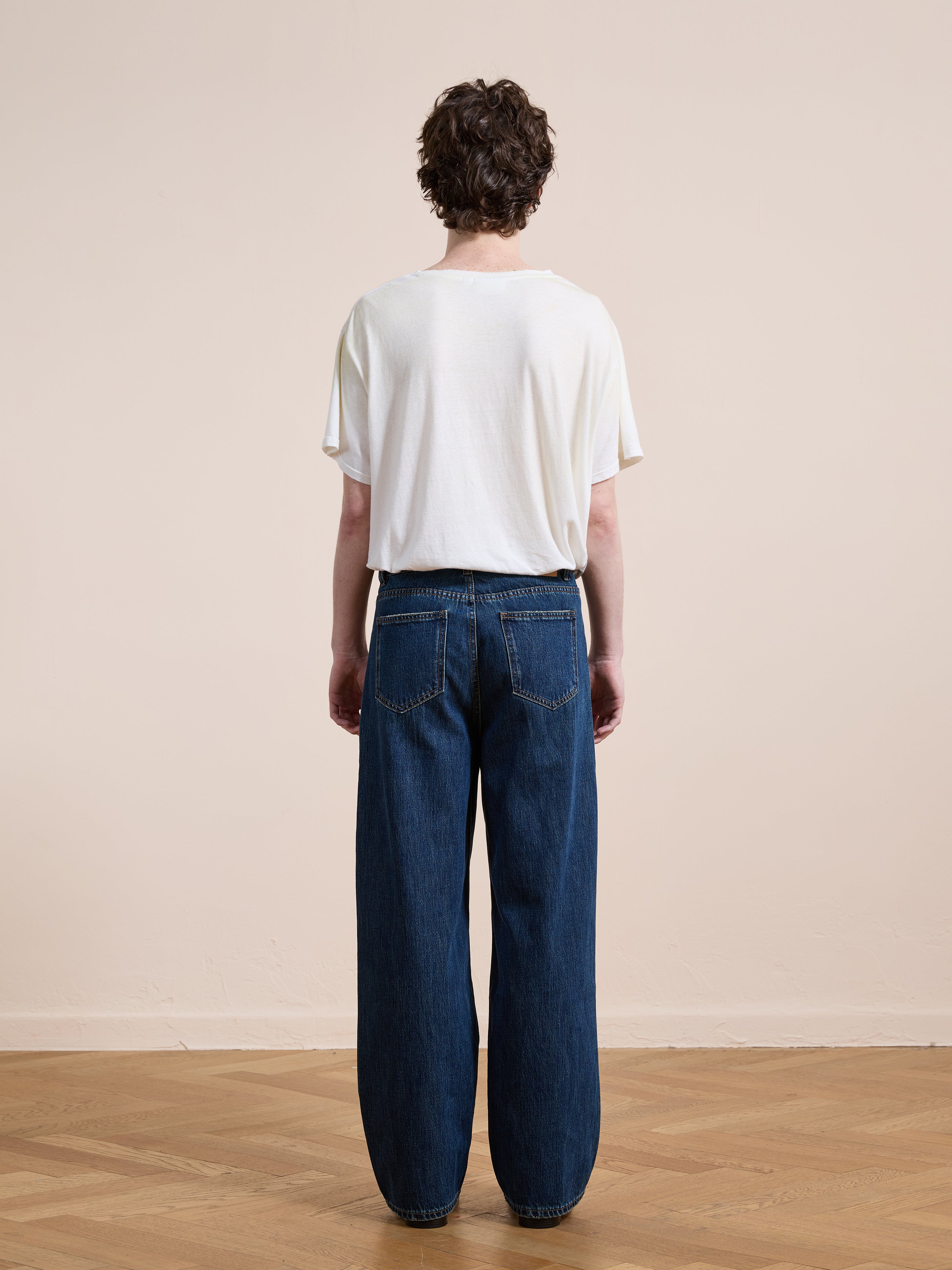 A person stands on a wooden floor facing away from the camera, wearing a loose white t-shirt and Found Pleated Relaxed Fit Jeans.