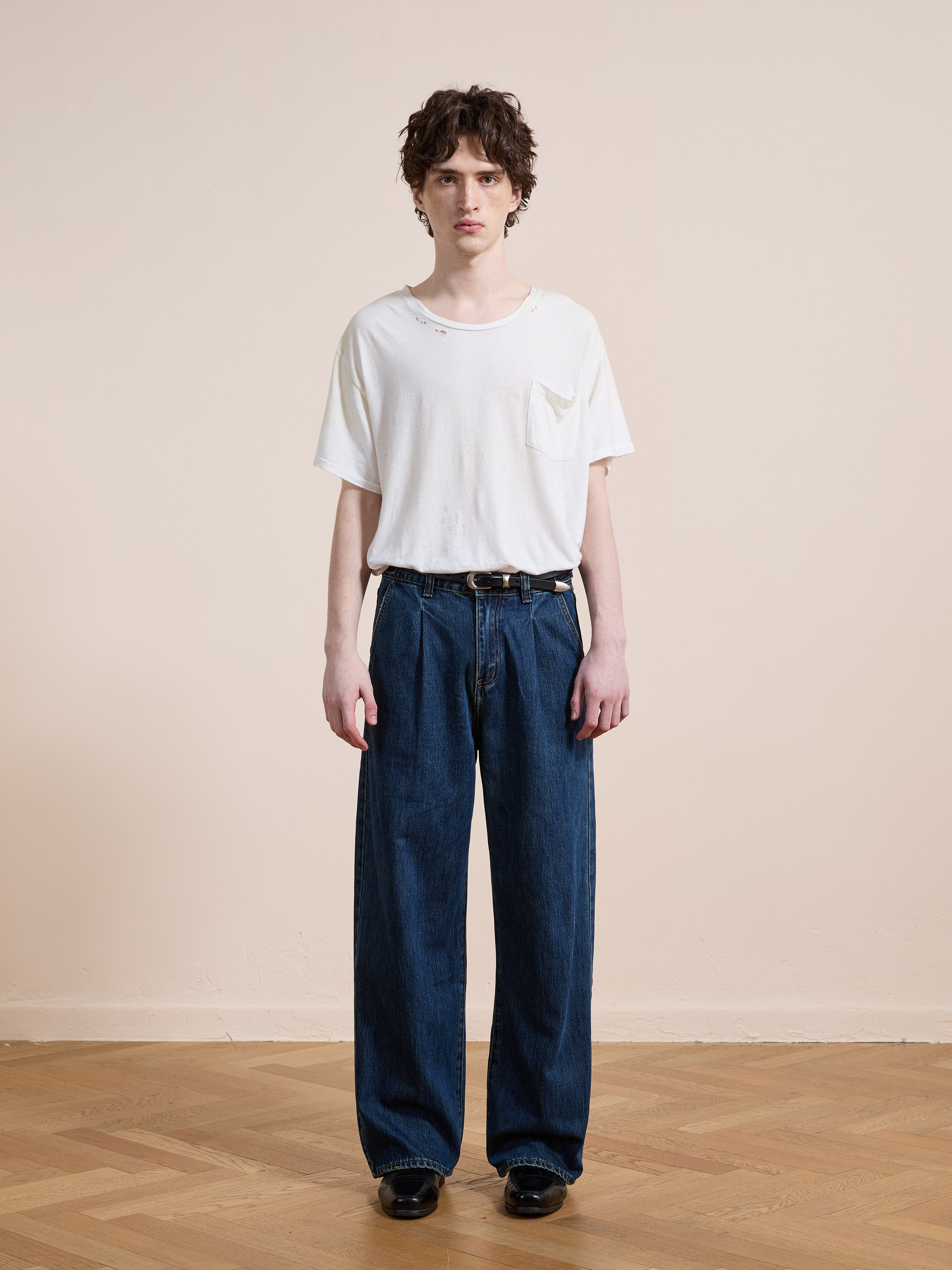 A person stands indoors on a wooden floor, wearing a white t-shirt, Found Pleated Relaxed Fit Jeans in vintage denim, and black shoes, facing the camera against a plain beige wall.