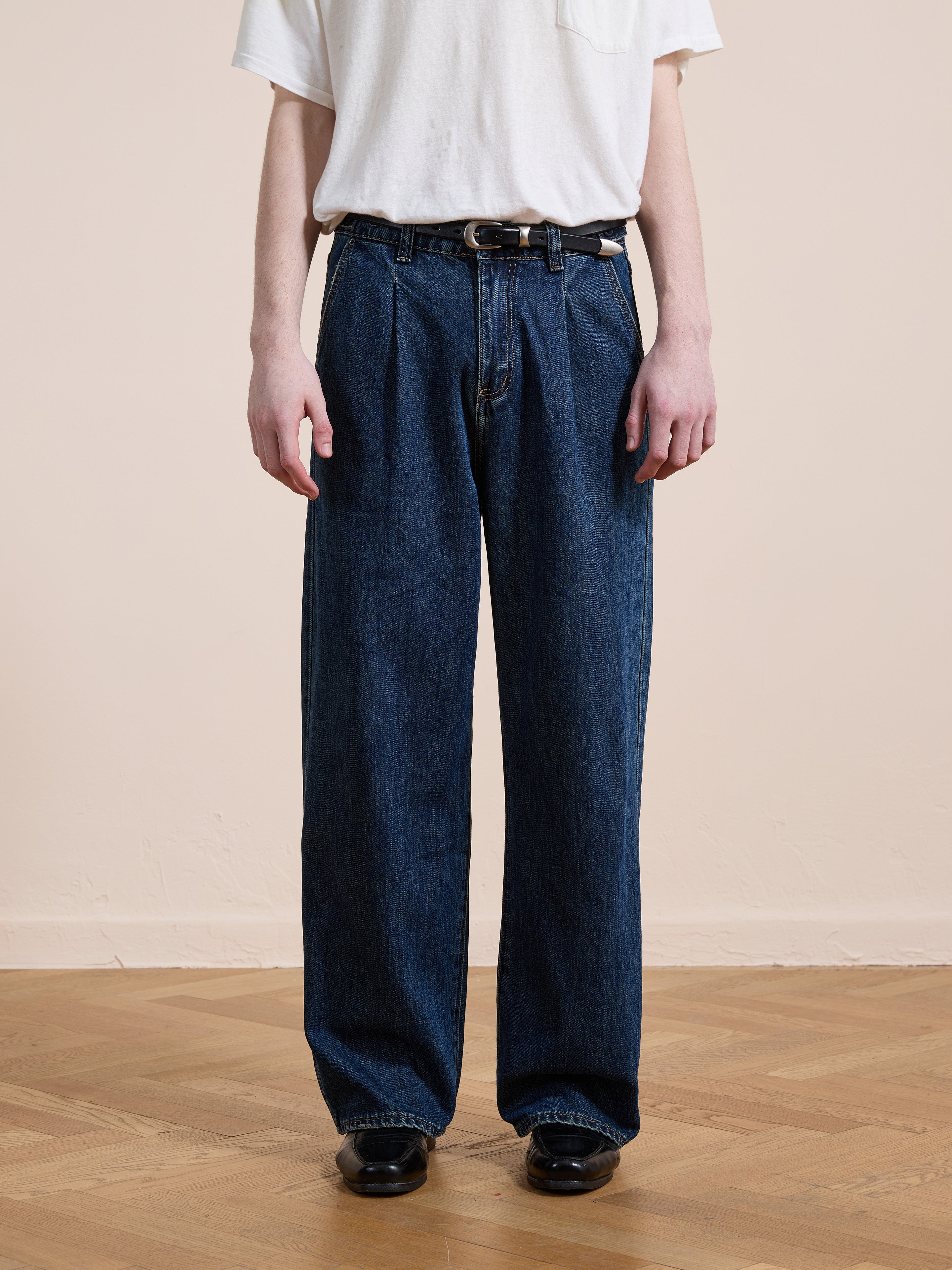 A person stands on a wooden floor against a plain beige background, wearing Found's Pleated Relaxed Fit Jeans in vintage denim with a belt, paired with a white T-shirt and black shoes.