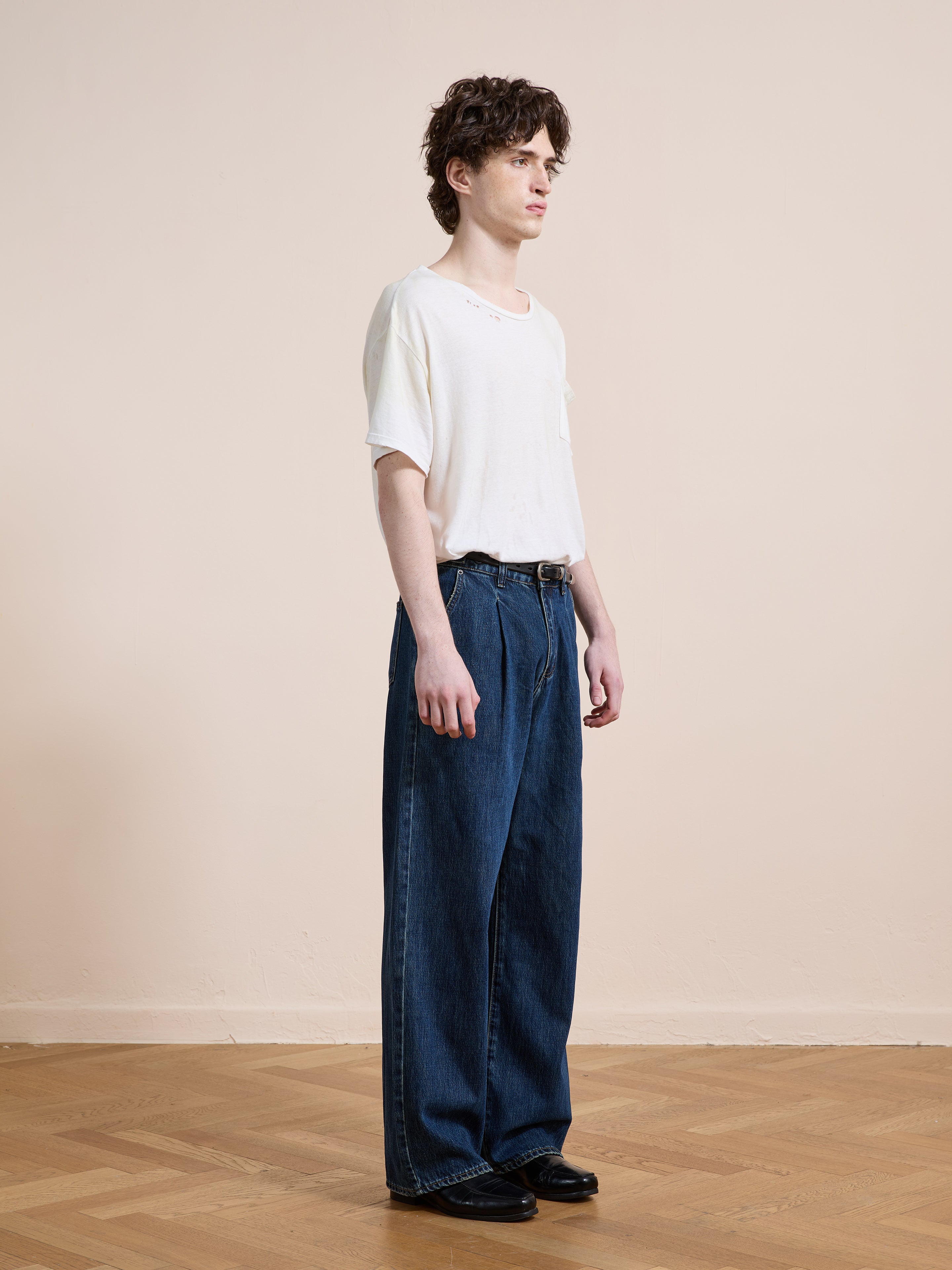 A person stands indoors on a wooden floor, wearing a white t-shirt, Found Pleated Relaxed Fit Jeans in vintage denim, and black shoes, facing slightly to the left against a plain beige wall.
