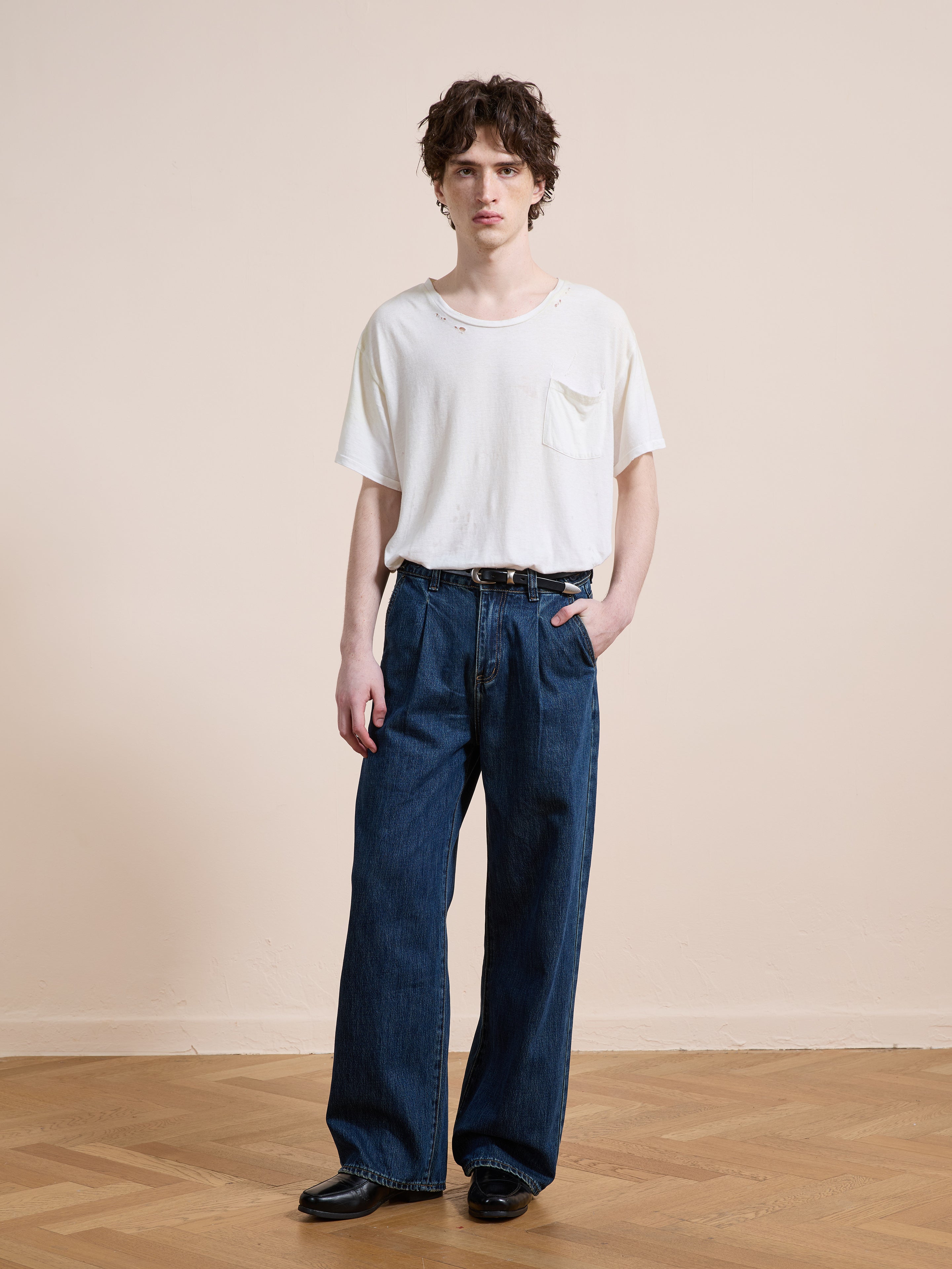 A person stands indoors on a wooden floor, wearing a loose white pocket t-shirt, Found Pleated Relaxed Fit Jeans in vintage denim, and black shoes, with one hand in their pocket against a plain beige wall.