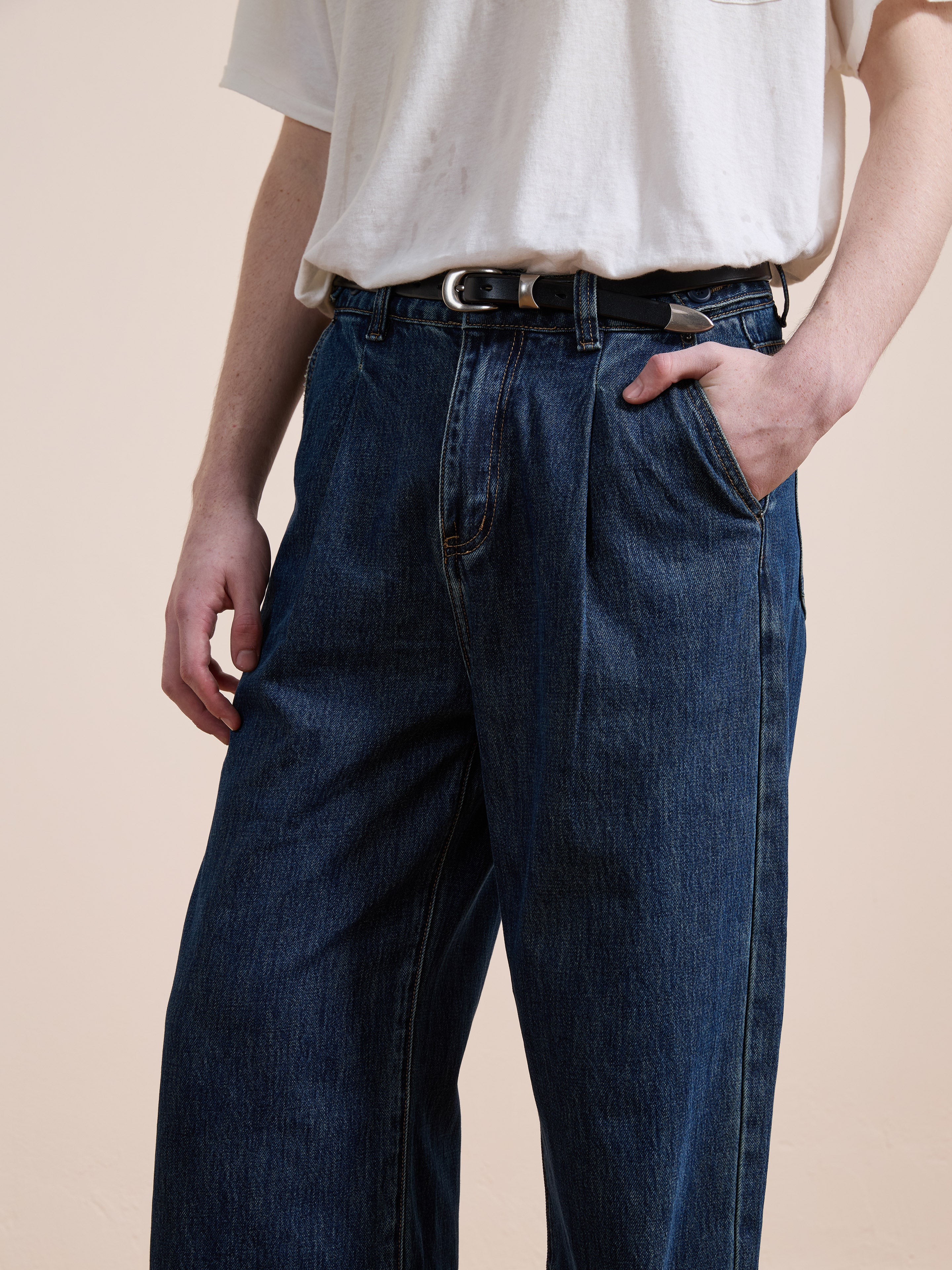 A person stands against a plain beige background wearing Found Pleated Relaxed Fit Jeans in dark blue, a white t-shirt, and a black belt, with one hand in their pocket.