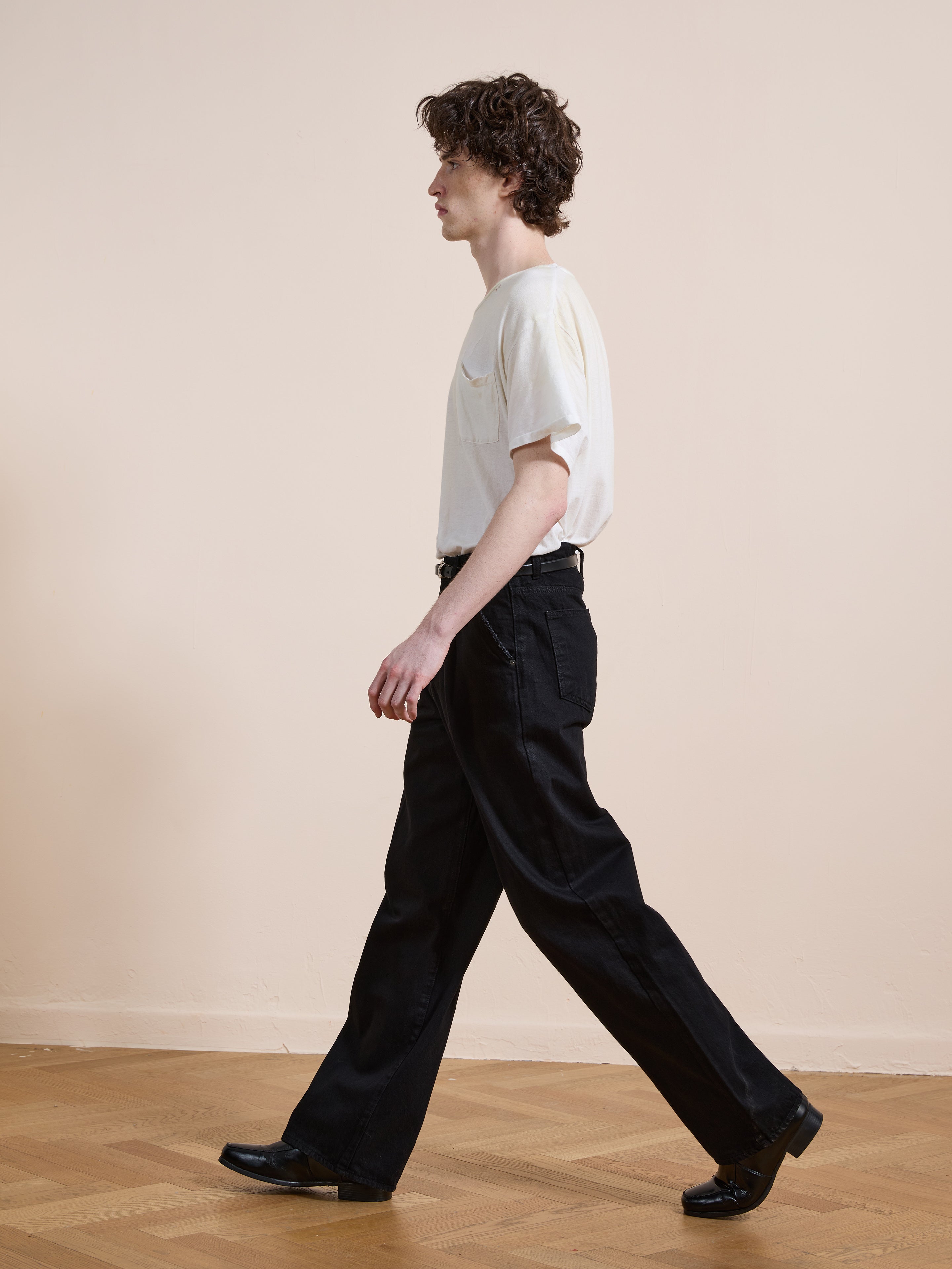 A person with curly hair walks indoors on a wooden floor, wearing a white t-shirt, Found Straight Leg Jeans, and black shoes against a plain beige wall.